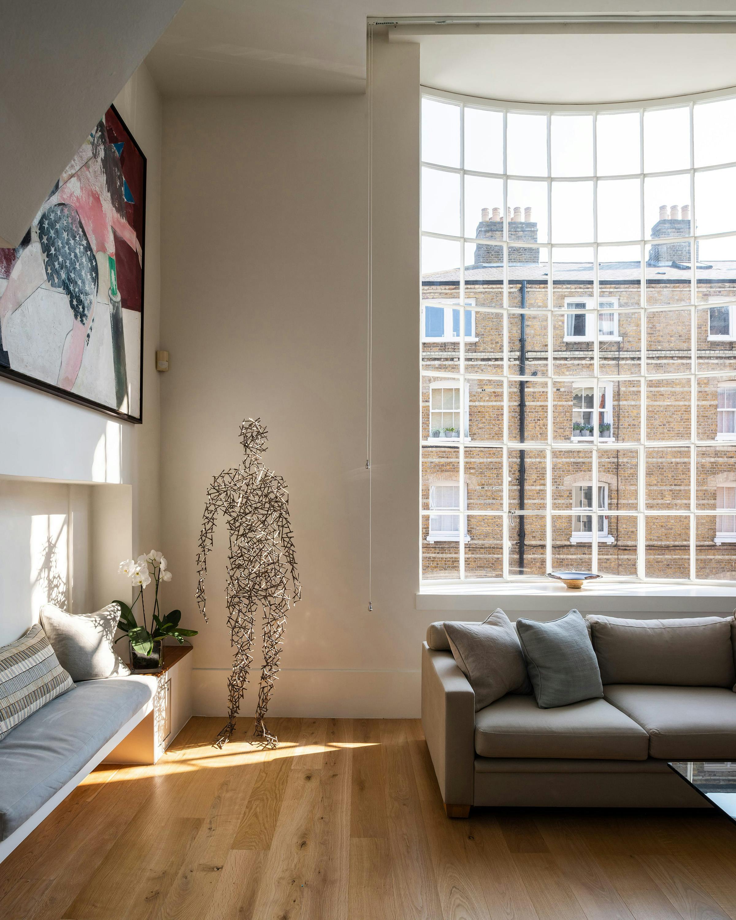 nterior shot of a bright living room in a London apartment. A large, arched window with multiple panes overlooks a traditional brick building with Georgian sash windows. A wire figure sculpture stands in the room, along with a modern sofa, wooden floors, and a colorful abstract painting.
