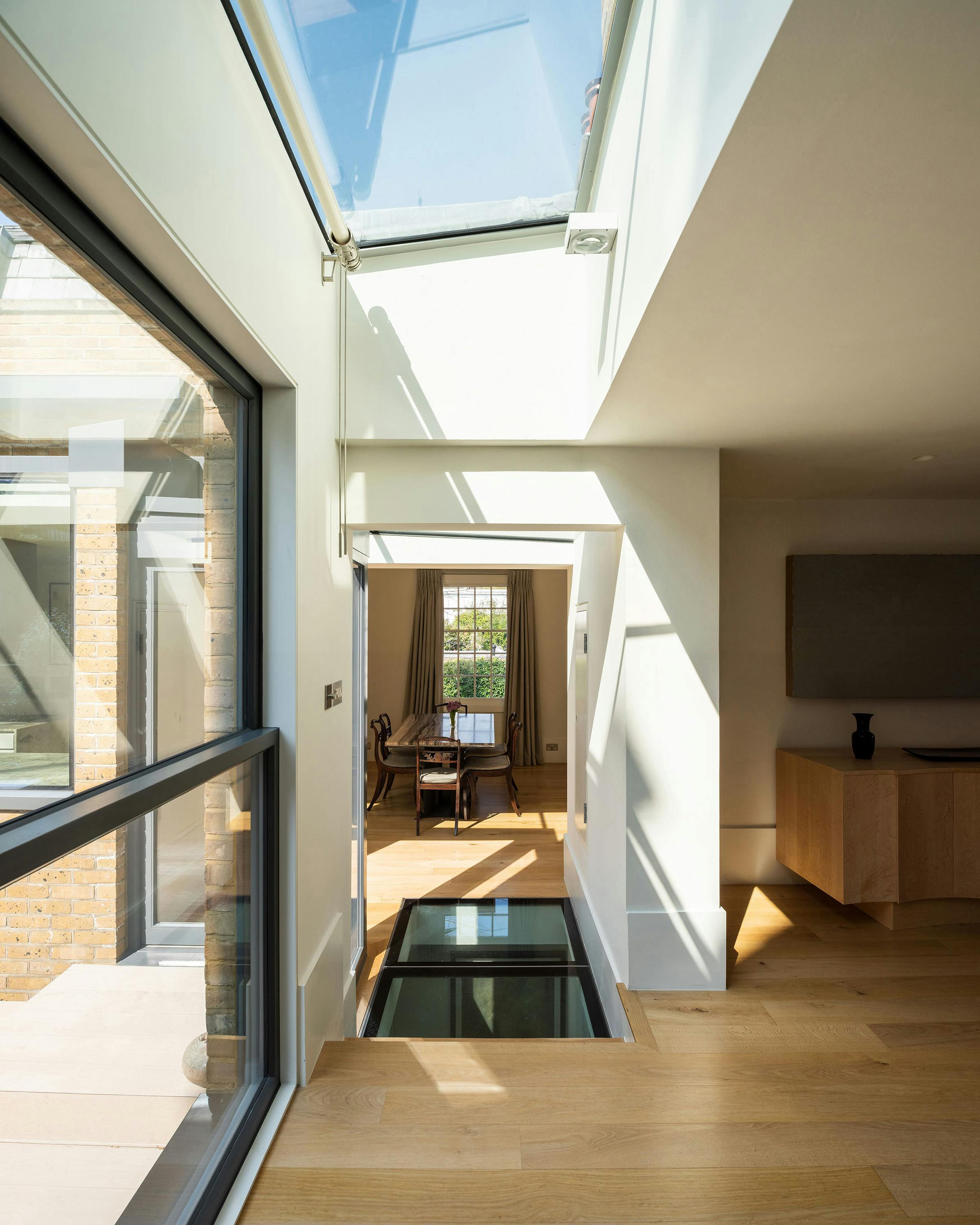 An interior shot of a modern hallway with light wood flooring. A glass panel in the floor reveals a space below. A large skylight illuminates the space. A doorway leads to another room with a dining table and windows. A glimpse of brickwork is visible through a large window on the left.