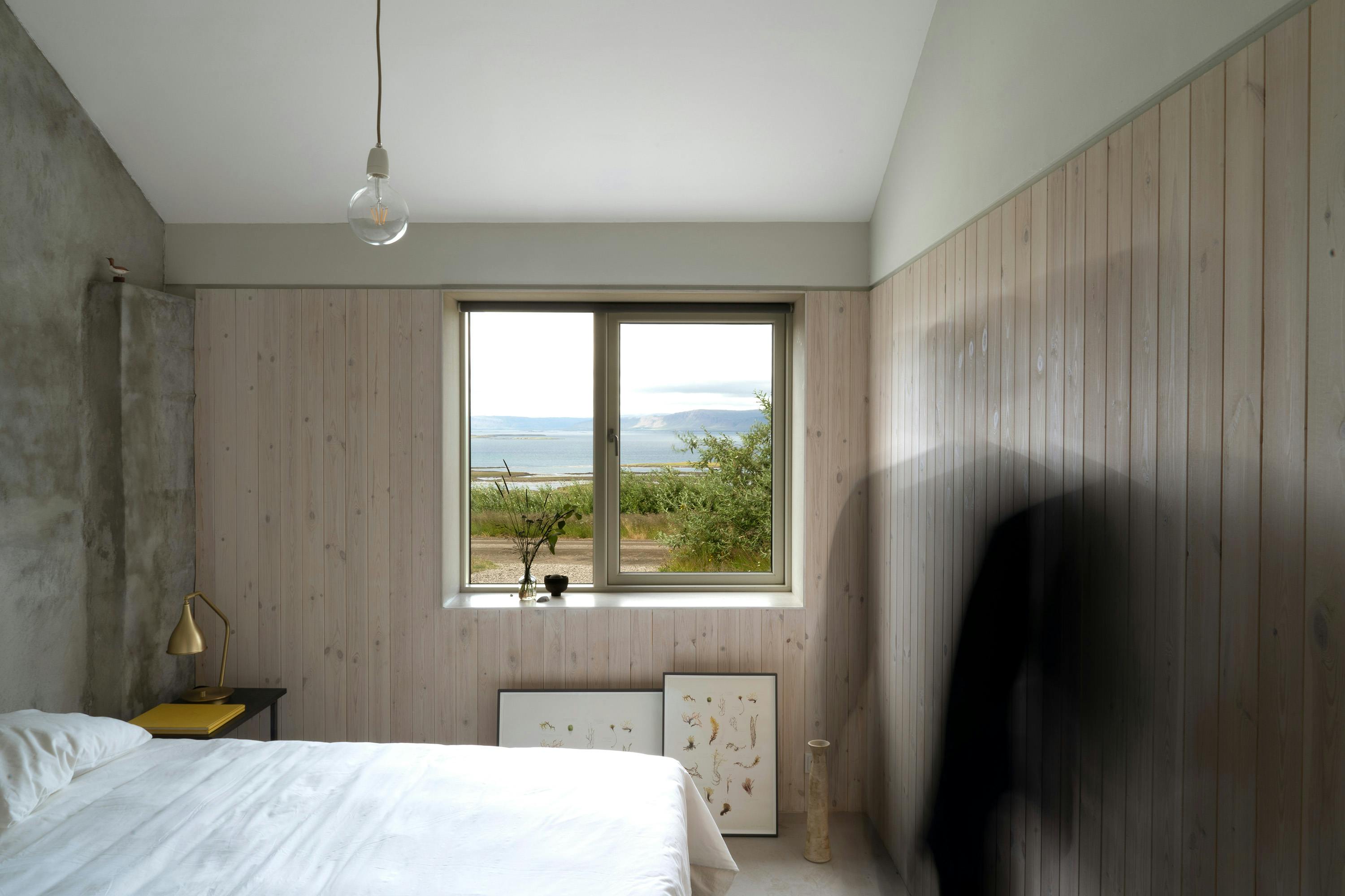 The image shows a bright, minimalist bedroom with a large window overlooking a scenic outdoor view