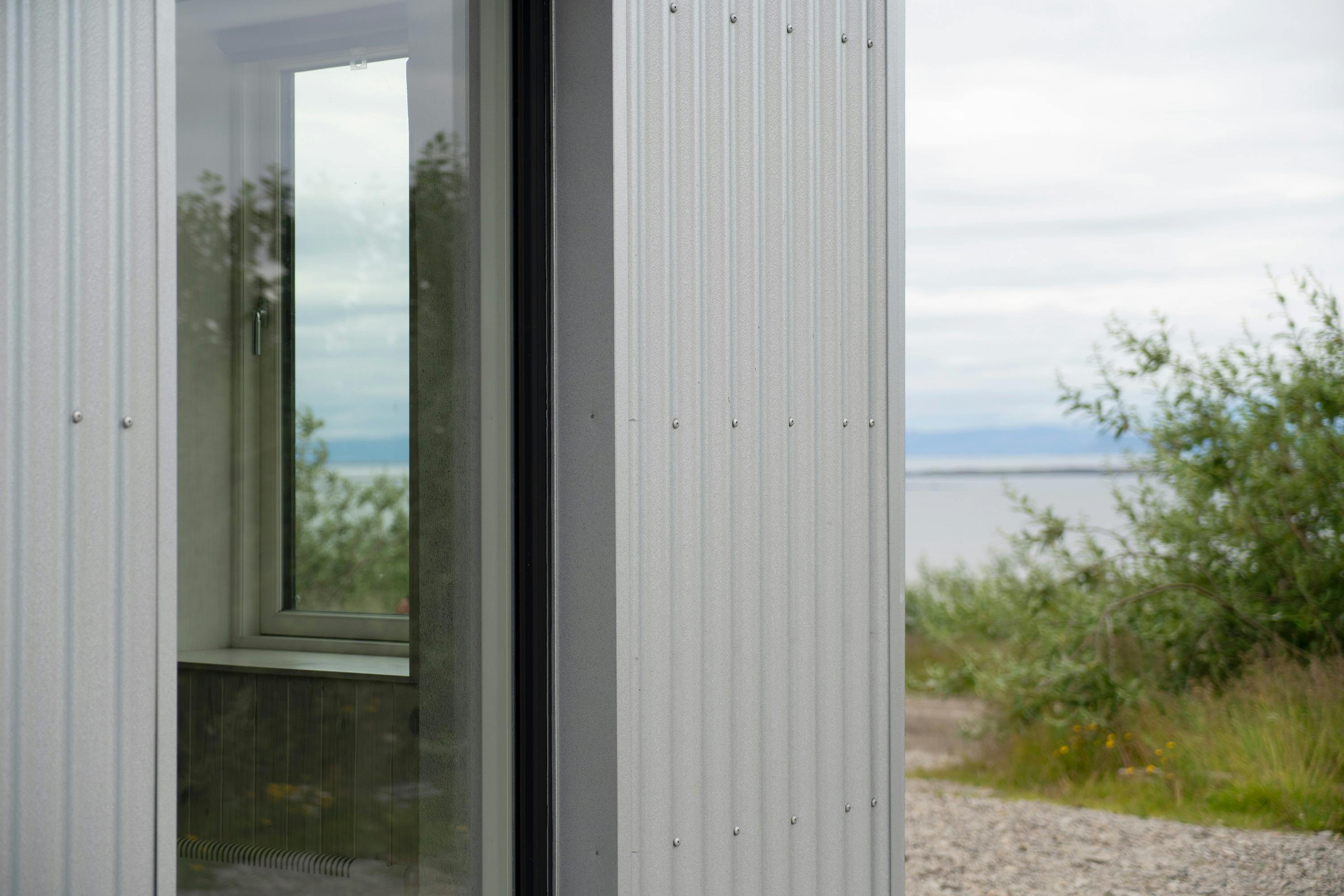 A close-up, angled shot of the exterior of a modern building with corrugated metal siding in a light gray or white color. The siding features evenly spaced rivets. A narrow, vertical window with a dark frame is visible, reflecting the outdoor scenery. Through the window, a glimpse of the interior shows a light-colored sill. The background reveals a gravel path leading to a body of water, possibly a lake or ocean, with a distant shoreline and a cloudy sky. Green shrubs and foliage are visible in the foreground, creating a natural contrast with the industrial feel of the building's siding. The shot is taken during daylight hours, but the overall lighting is soft and diffused.
