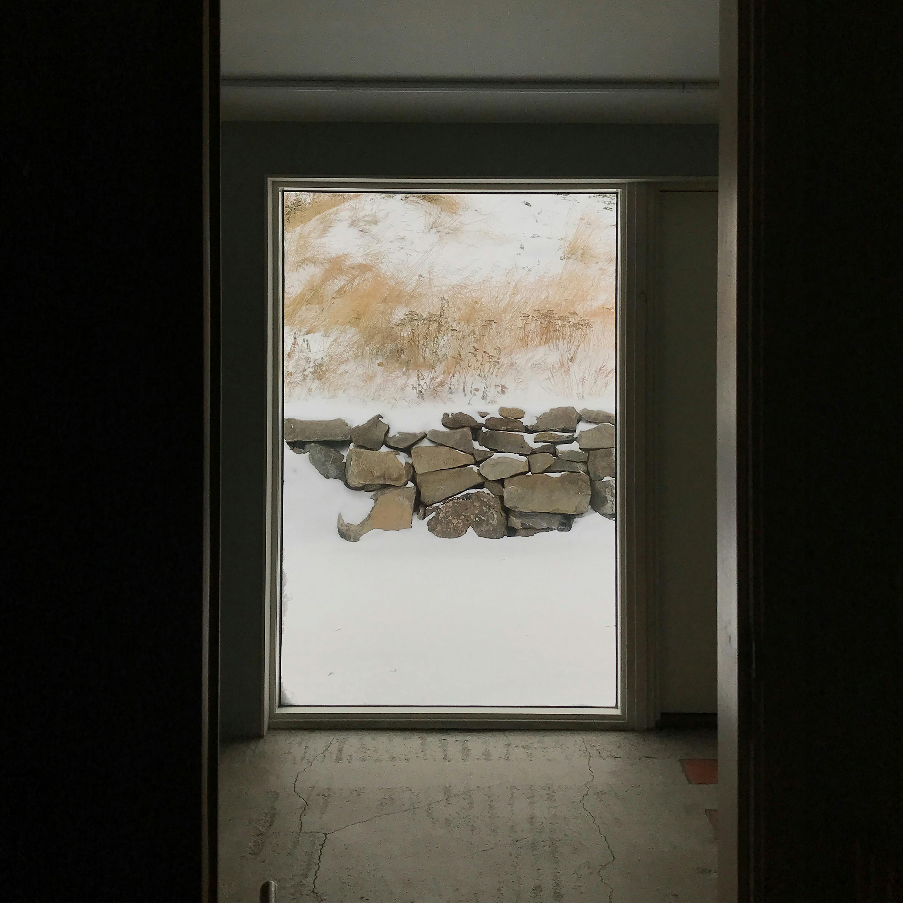 A slightly angled shot through a doorway into a room with a large rectangular window at the far end. The doorway is framed by dark door jambs, creating a perspective view into the room. The room's floor is a light, cracked concrete or stone, suggesting an unfinished or rustic space. The large window at the end of the room shows an outdoor scene. The scene features a low stone wall at the bottom, partially covered in snow. Above the wall, the ground is a mix of brown, dry grass and patches of snow. The sky is overcast and appears to be a light gray, suggesting a winter day. The window frame is thin and light-colored, contrasting with the dark doorway. The overall image has a slightly muted and desaturated color palette, emphasizing the rustic and wintry feel of the scene.