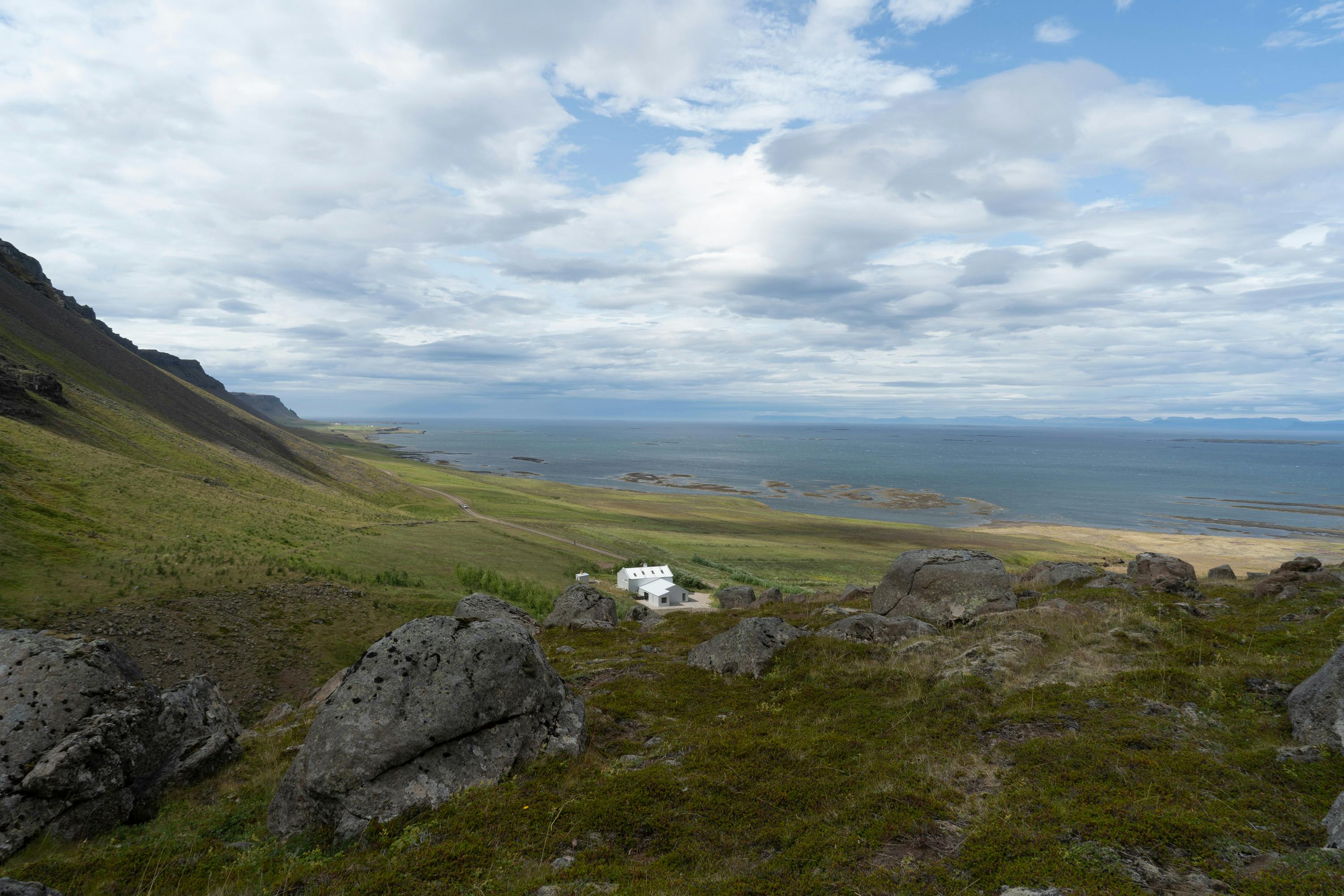 A wide, scenic landscape shot of a coastal area. The foreground features large, dark, lichen-covered rocks scattered across a grassy hillside. In the midground, a white building with a gray roof sits nestled in a green valley, with a faint path leading towards it. The valley slopes down to meet a wide expanse of water, possibly a bay or ocean, which extends to the horizon. The water is a deep blue-gray color. In the distance, a faint coastline is visible. The sky is filled with white, fluffy clouds against a light blue backdrop, suggesting a partly cloudy day. The overall scene conveys a sense of remote tranquility and natural beauty.