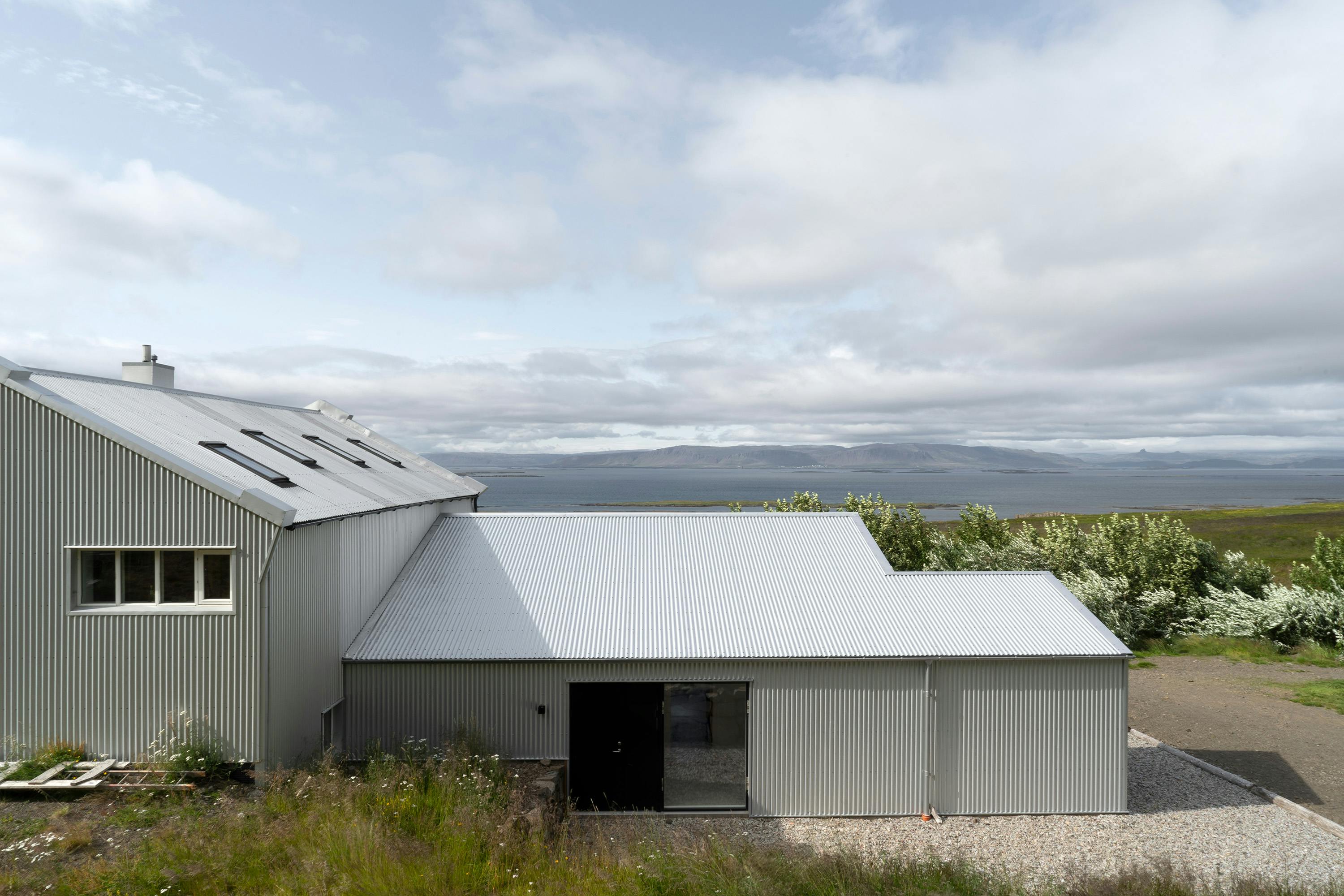 An eye-level, slightly angled shot of a modern building with corrugated metal siding. The building consists of two interconnected sections. The larger section on the left has a sloped roof with several windows. The smaller section on the right has a flatter, lower roof and a large, dark glass door. Both sections are clad in light gray or white corrugated metal. The ground in front of the building is a mix of gravel and patches of grass. In the background, a body of water, possibly a lake or ocean, stretches to the horizon with a distant shoreline and a cloudy sky. The overall scene conveys a contemporary architectural style in a natural, outdoor setting.