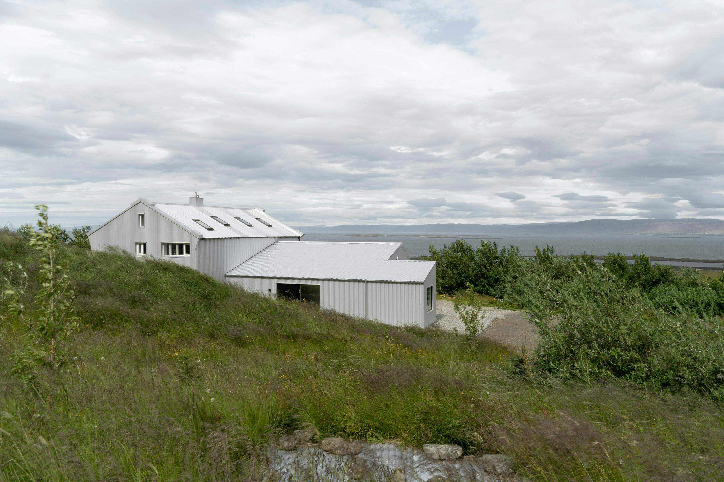 A wide-angle, slightly elevated shot of a modern building with corrugated metal siding, situated on a grassy hillside. The building consists of two interconnected sections. The larger section on the left has a sloped roof with several small, rectangular windows. The smaller section on the right has a flatter, lower roof and a dark glass door. Both sections are clad in light gray or white corrugated metal. The hillside slopes down from the foreground towards the building, with tall, wild grasses and foliage dominating the scene. A rocky outcropping is visible at the bottom of the frame. In the background, a body of water, possibly a lake or ocean, stretches to the horizon with a distant shoreline and a cloudy sky. The overall scene conveys a sense of remote tranquility and contemporary architectural style integrated into a natural, outdoor setting.