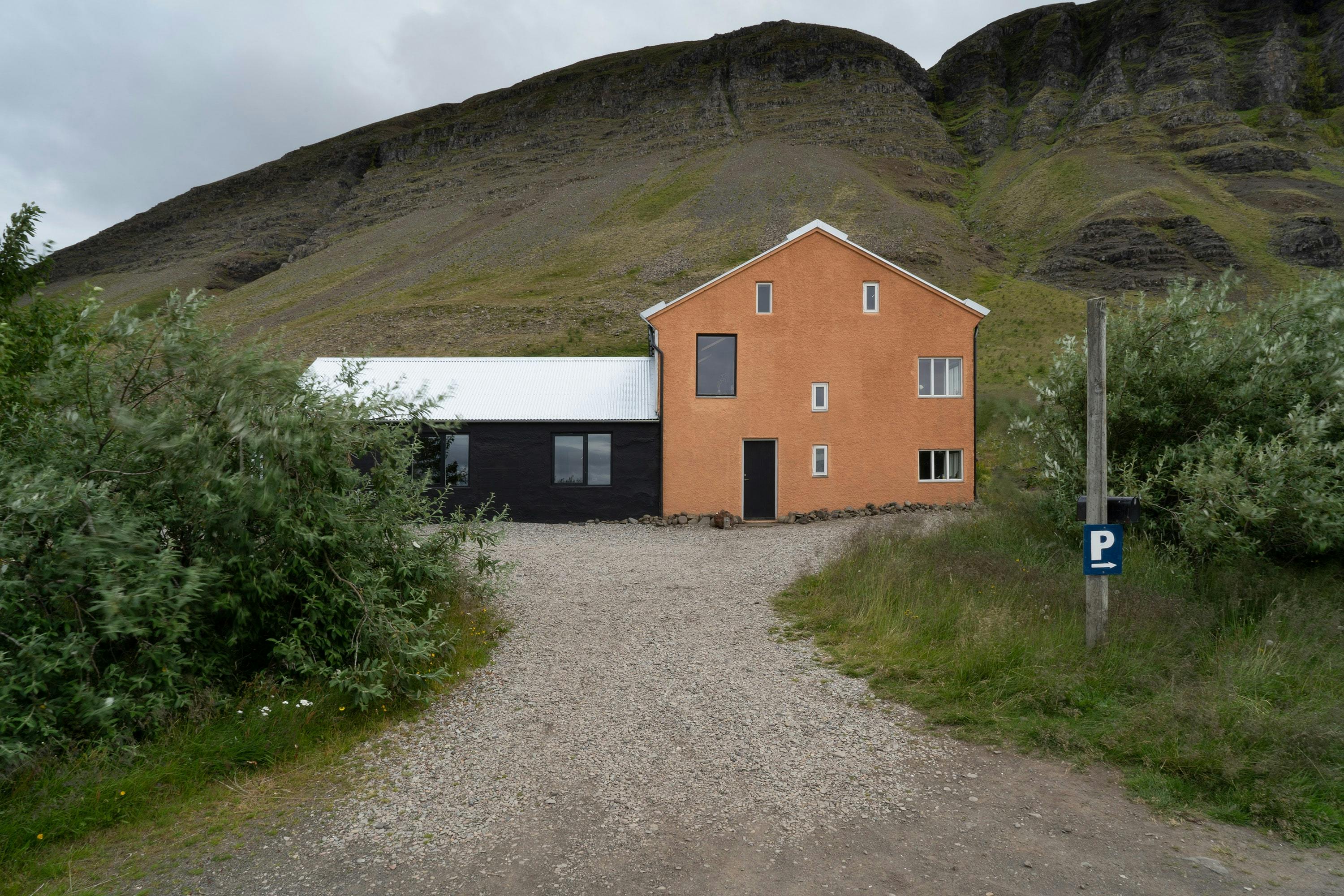 A straight-on shot of a two-section building with a striking color contrast, situated at the end of a gravel driveway. The building is composed of two distinct parts: the section on the left is dark, possibly black or dark gray, with a flat roof and a single window visible. The section on the right is a bright, peach or orange color with a pitched roof and several small, rectangular windows. A dark door is centered on the front of the orange section. The gravel driveway leads directly to the building, creating a central pathway in the image. Lush green bushes flank the driveway on both sides. In the background, a large, dark, rocky hill or mountain rises, dominating the upper portion of the frame. A blue sign with the letter "P" is visible on a post in the right foreground. The sky is overcast and gray. The image conveys a sense of a remote or secluded dwelling with a unique architectural style.