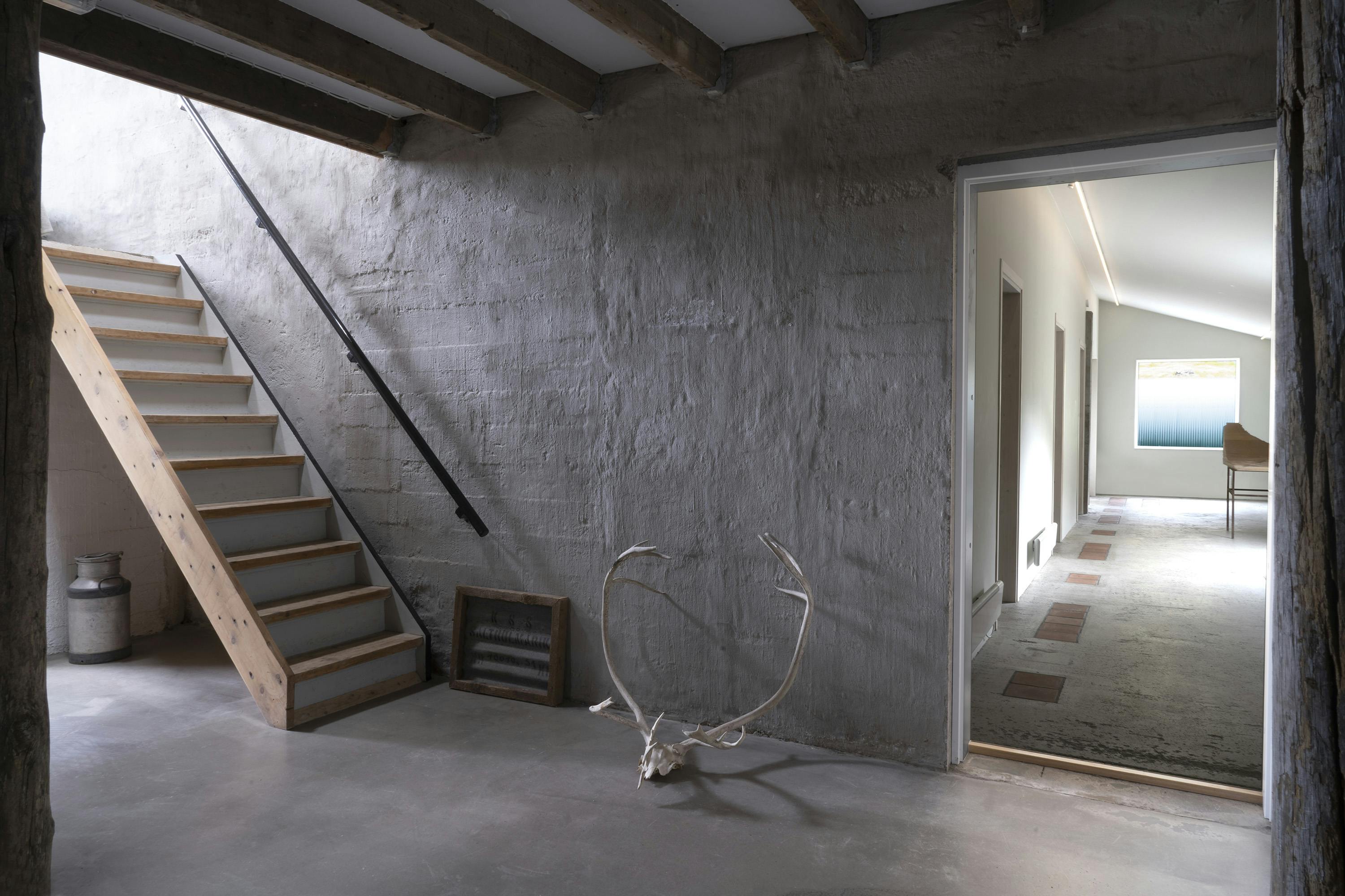 A slightly angled shot of an interior space with a rustic, unfinished aesthetic. The space features a concrete floor and textured, gray plastered walls. A wooden staircase with open risers and a black metal handrail is visible on the left, leading upwards. To the right, an open doorway reveals a hallway with a sloped ceiling, suggesting an attic conversion. The hallway has light-colored walls and a patterned floor. In the foreground, a large, antler-like object rests on the concrete floor, along with a metal container and a framed object with a grid pattern. The ceiling has exposed wooden beams. The lighting is soft and diffused, highlighting the textures of the walls and floor. The overall scene conveys a sense of raw, natural materials and a minimalist design.