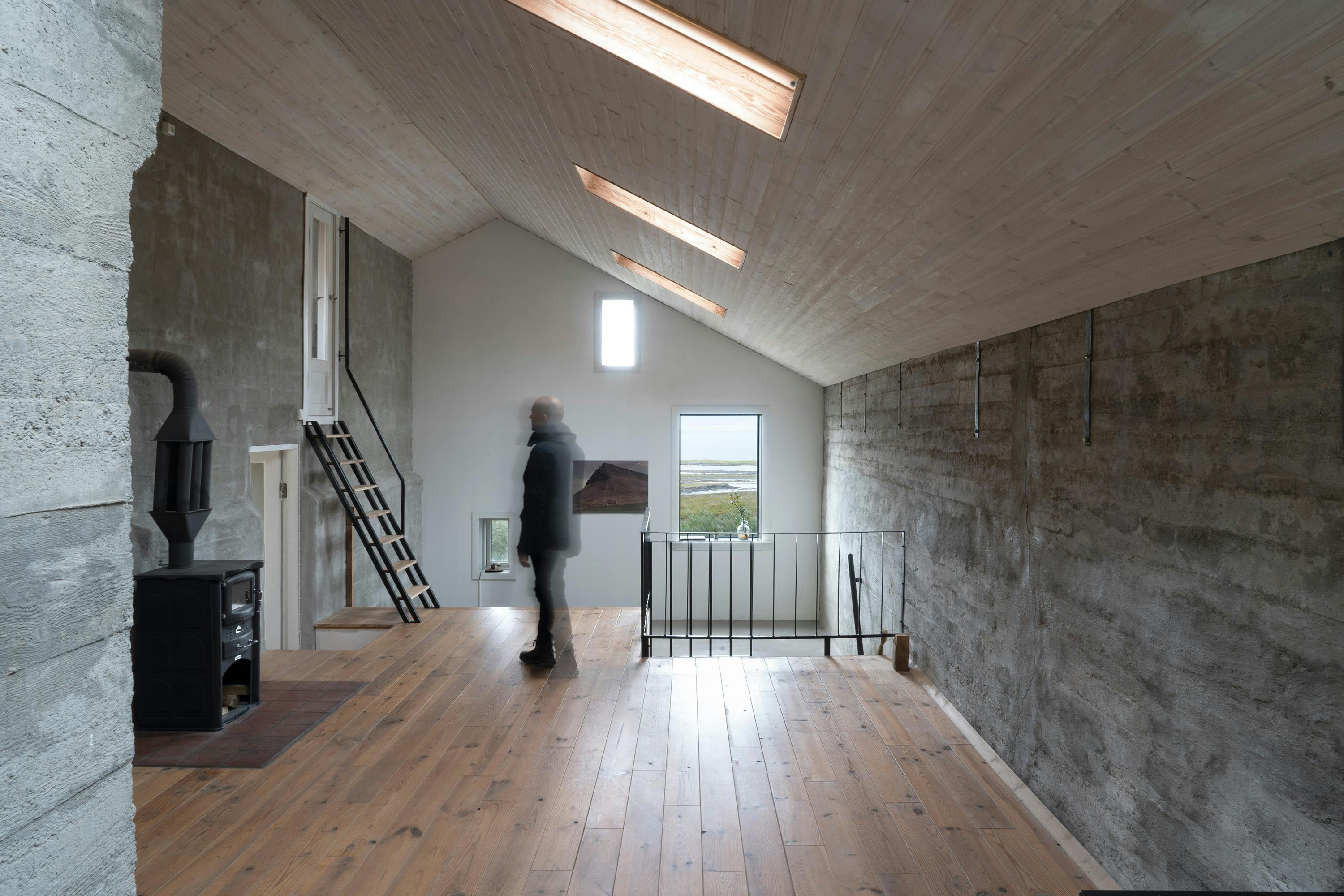 An interior shot of a spacious room with a minimalist design, featuring a person standing in the center. The room has a high, sloping ceiling with three rectangular skylights that emit a warm light. The walls are made of textured, gray concrete, creating a raw and industrial feel. The floor is constructed of wide, light-colored wooden planks. On the left side, a black wood-burning stove is visible, along with a metal staircase leading to an upper level. A doorway is also present, leading to another room. A person wearing a dark coat is standing in the center of the room, facing away from the viewer. On the right, a black metal railing separates the room from a lower level or another space. A window at the far end of the room offers a view of an outdoor landscape with a body of water and green vegetation. The lighting is a mix of natural light from the skylights and the window, creating a bright and airy atmosphere. The overall scene conveys a sense of modern, minimalist architecture with a focus on natural materials and light.