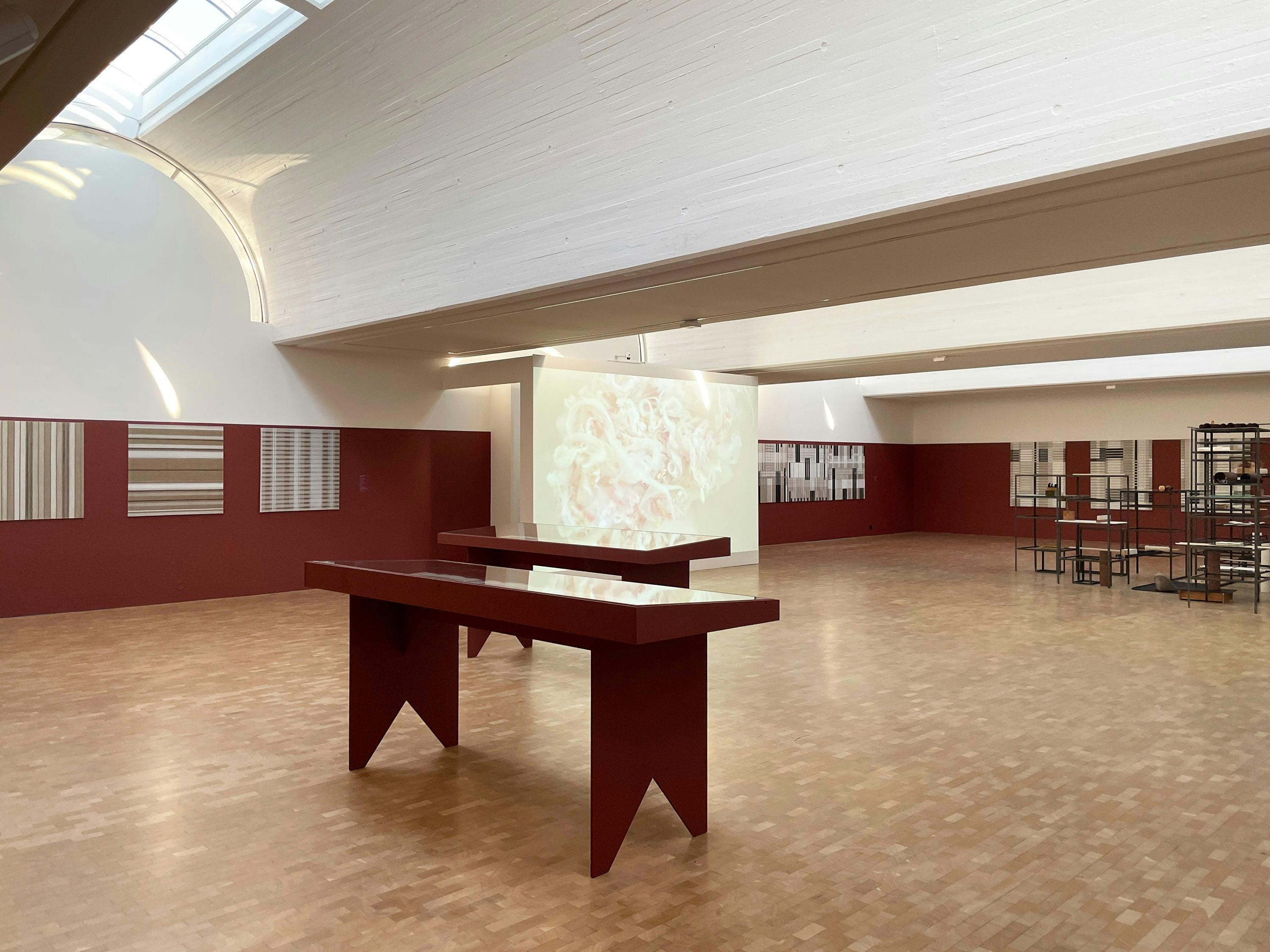 A slightly elevated, straight-on photograph of a large, well-lit room with an art installation. The room features light-coloured walls and a herringbone patterned wood floor. On the left and right sides of the room, there are red-painted display walls with framed artworks featuring striped patterns. In the centre of the room, two long, red-painted tables with a simple, angular design are placed. Behind the tables, a large screen or wall projection displays a blurry, light-coloured image. On the right side of the room, a metal structure with multiple shelves is visible. The room has a high, vaulted ceiling with skylights, providing natural light. The overall aesthetic is modern and minimalist, with a focus on clean lines and geometric shapes.