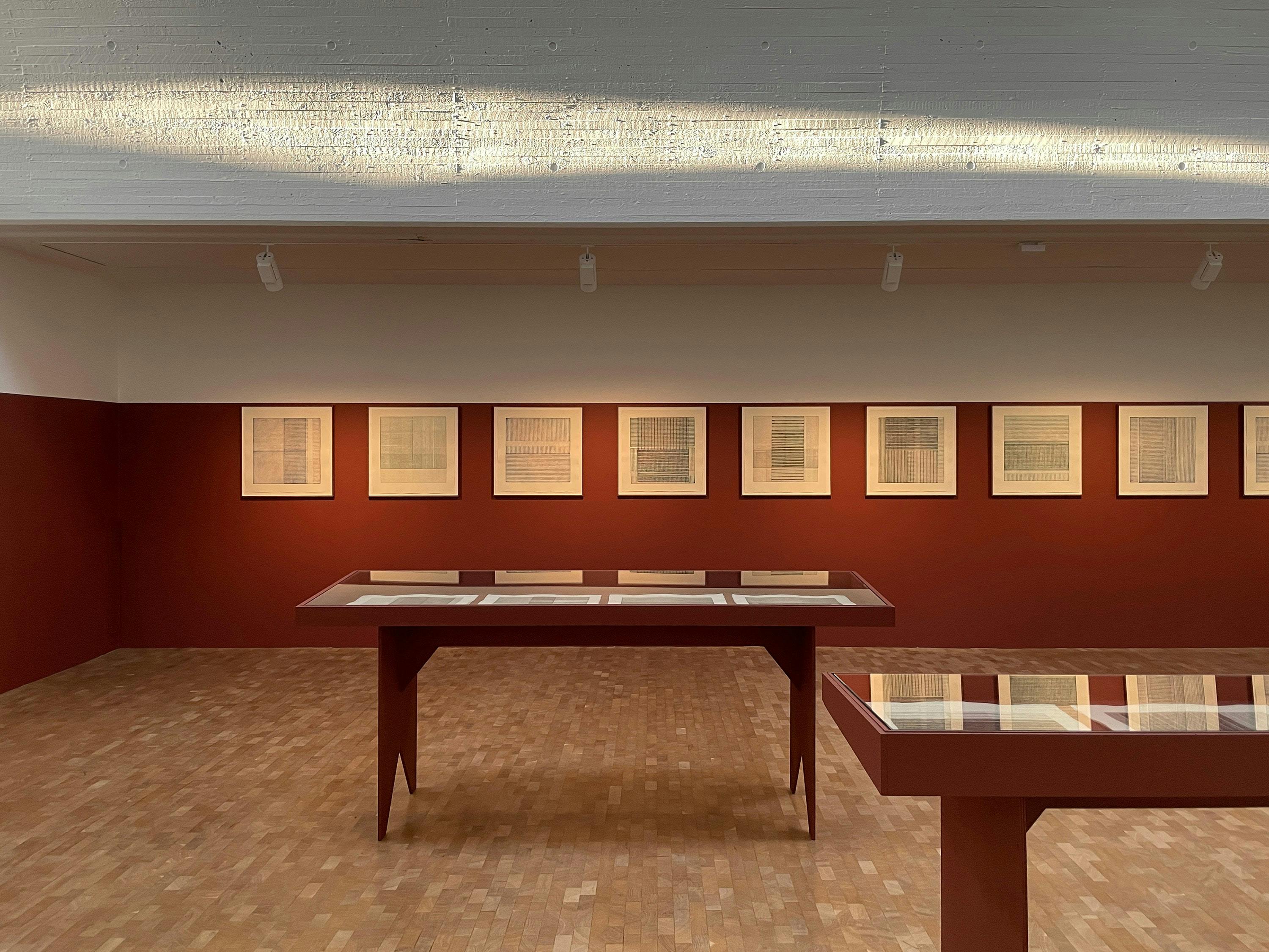 A slightly elevated, straight-on photograph of an art gallery space. The room features a light-coloured ceiling with visible texture, and a herringbone patterned wood floor. A red-painted wall runs along the back of the room, featuring a series of framed artworks with grid-like patterns. In the centre of the room, two long, red-painted tables with a simple, angular design are placed. The tables have glass tops, and similar grid-like patterns are visible beneath the glass. The ceiling has recessed lighting fixtures. Sunlight casts a warm glow on the ceiling, suggesting a window or skylight out of frame. The overall aesthetic is modern and minimalist, with a focus on clean lines and geometric shapes.