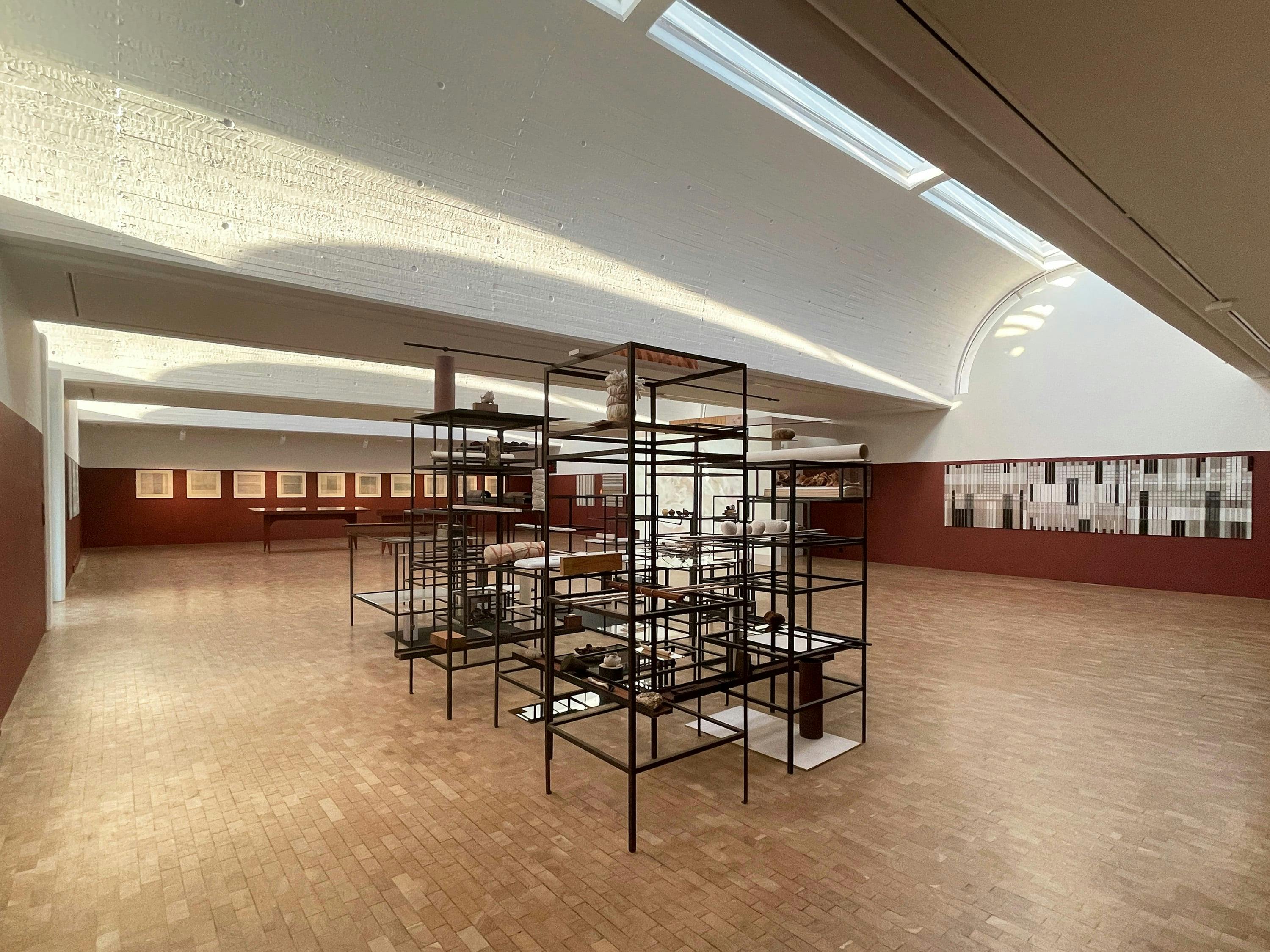 A slightly elevated, straight-on photograph of a large, well-lit room with an art installation. The room features light-coloured walls, a vaulted ceiling with skylights, and a herringbone patterned wood floor. In the centre of the room, a large, multi-tiered metal structure with shelves is placed. The structure is black and features various small objects and displays on the shelves. On the left and right sides of the room, there are red-painted display walls with framed artworks featuring grid-like patterns. Sunlight casts a warm glow on the ceiling, highlighting its texture and the skylights. The overall aesthetic is modern and minimalist, with a focus on geometric shapes and natural light.