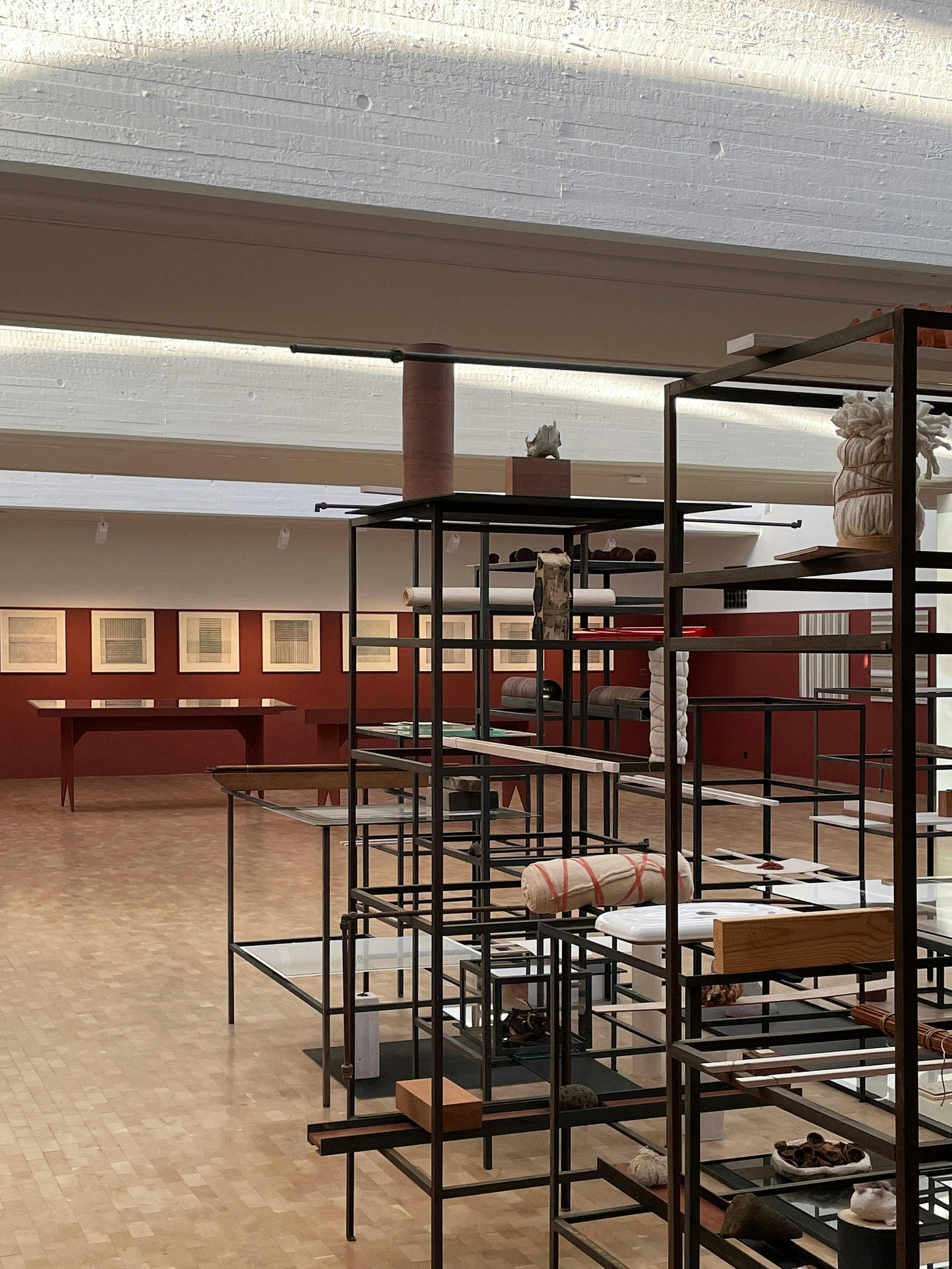 A slightly elevated, angled photograph of a large, well-lit room with an art installation. The room features a light-coloured ceiling with exposed beams and a herringbone patterned wood floor. In the foreground, a large, multi-tiered metal structure with shelves is visible. The structure is black and features various small objects and displays on the shelves, including fabric rolls, ceramic pieces, and shoes. In the background, a red-painted wall with framed artworks featuring grid-like patterns is visible. A long, red table is placed against the wall. The ceiling has visible texture and exposed beams. The overall aesthetic is modern and minimalist, with a focus on geometric shapes and the display of various objects.