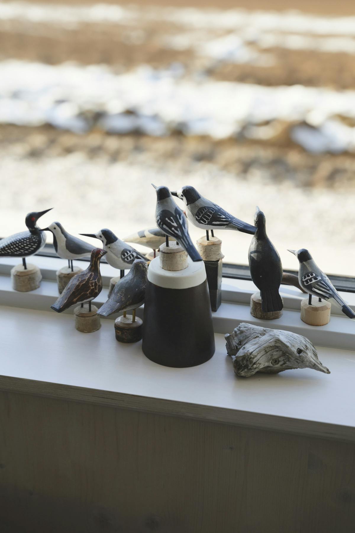 A collection of nine handcrafted wooden bird sculptures, including a loon, blue jays, and other species, are arranged on a bright white windowsill. A dark, cylindrical container with a light-colored top and a piece of weathered driftwood are also visible. A blurred snowy landscape is seen through the window.