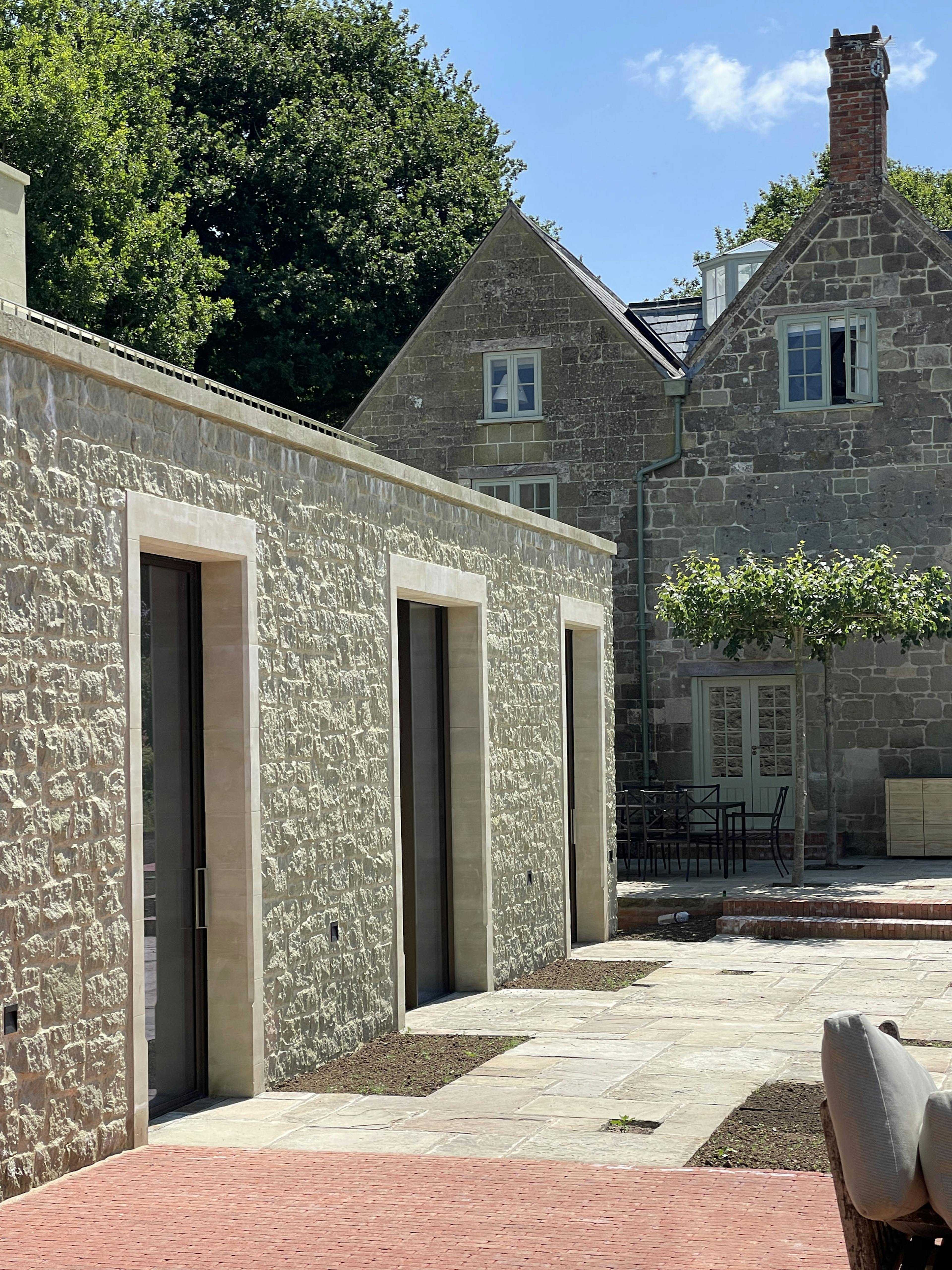A modern stone building in bright sun, with a terrace in red clay pavers. An older stone building in the background