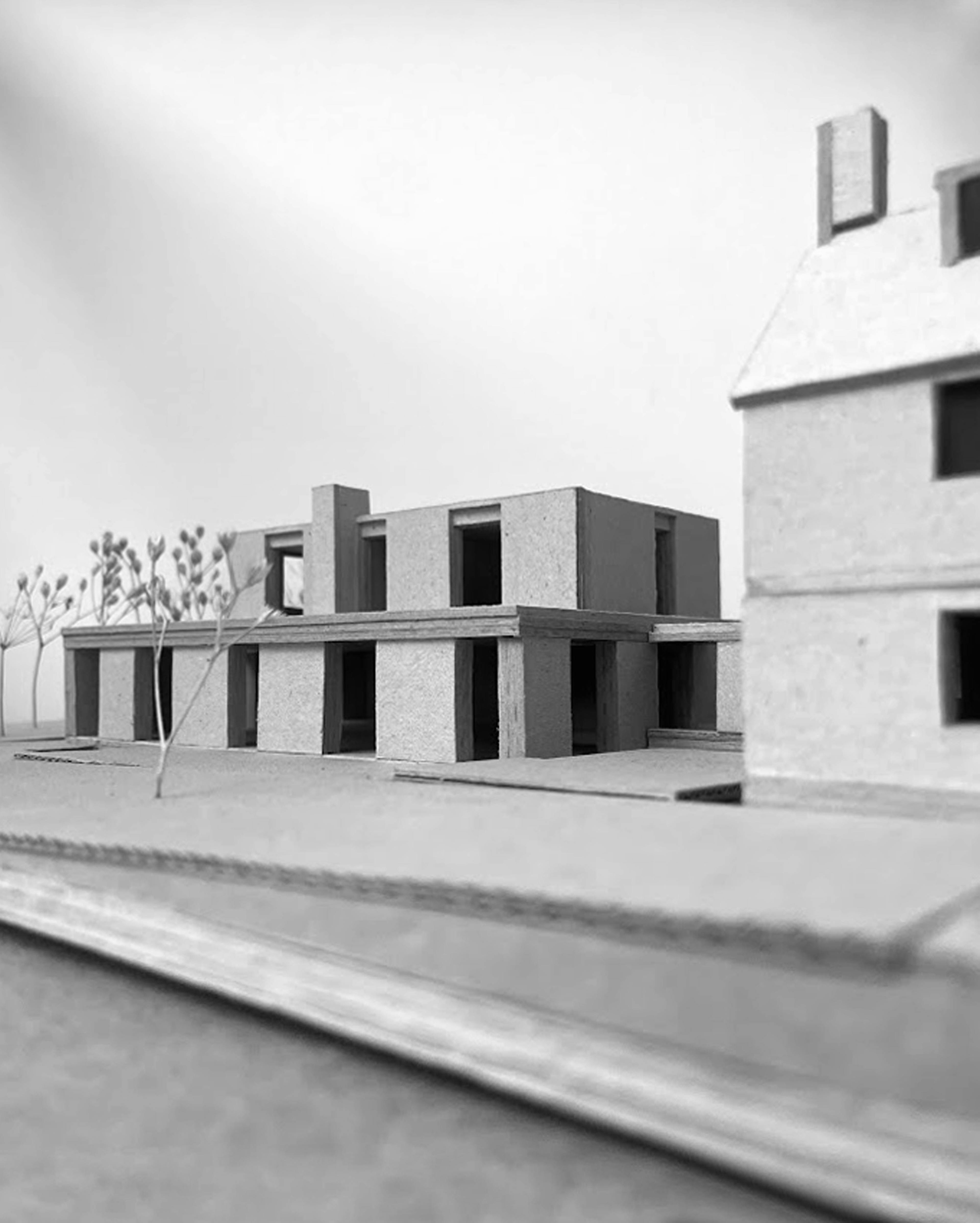 A detailed cardboard architectural model showcases a house with a pitched roof and a modern building on a raised platform with a retaining wall. Dried plant stems are used to represent trees and foliage.