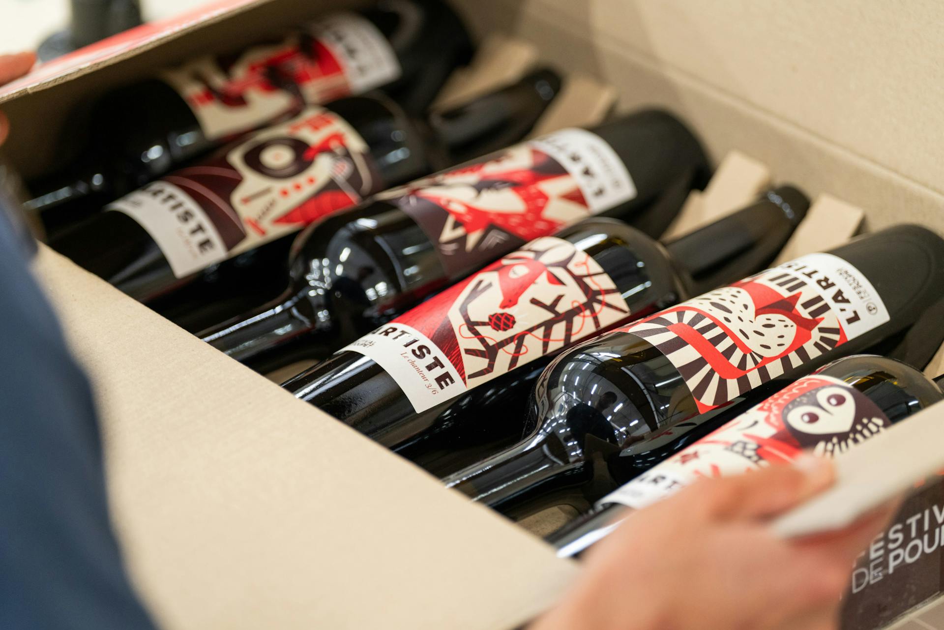 Une personne ouvrant un carton de bouteilles de vins de la cuvée du Festival de Poupet, arborant les étiquettes créés par l'agence de design Katra.