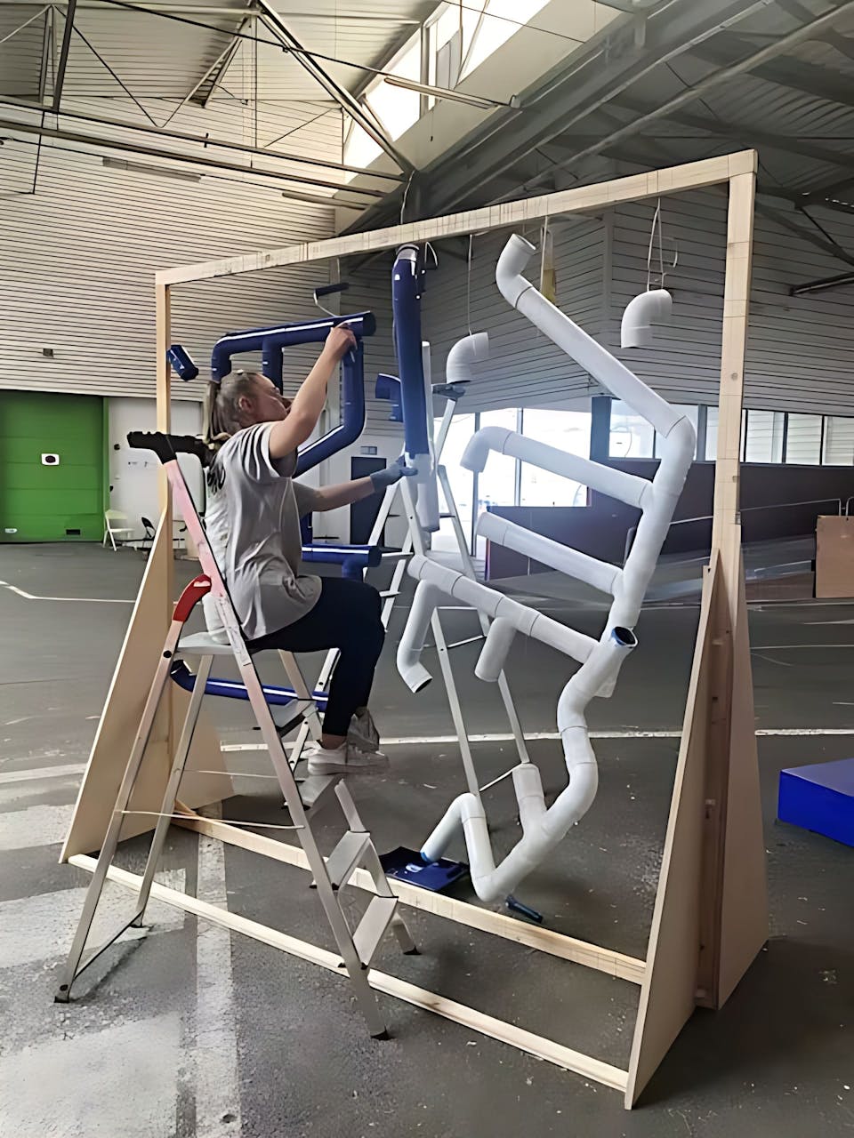 Phase de peinture sur un récupérateur d'eau en cours de fabrication. Une jeune femme sur un escabeau peint des tuyaux PVC.