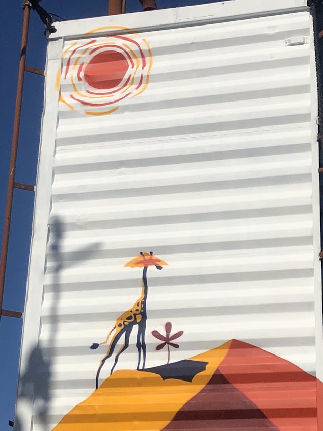 Une fresque murale extérieure sur un conteneur, représentant une girafe sous le soleil du désert.