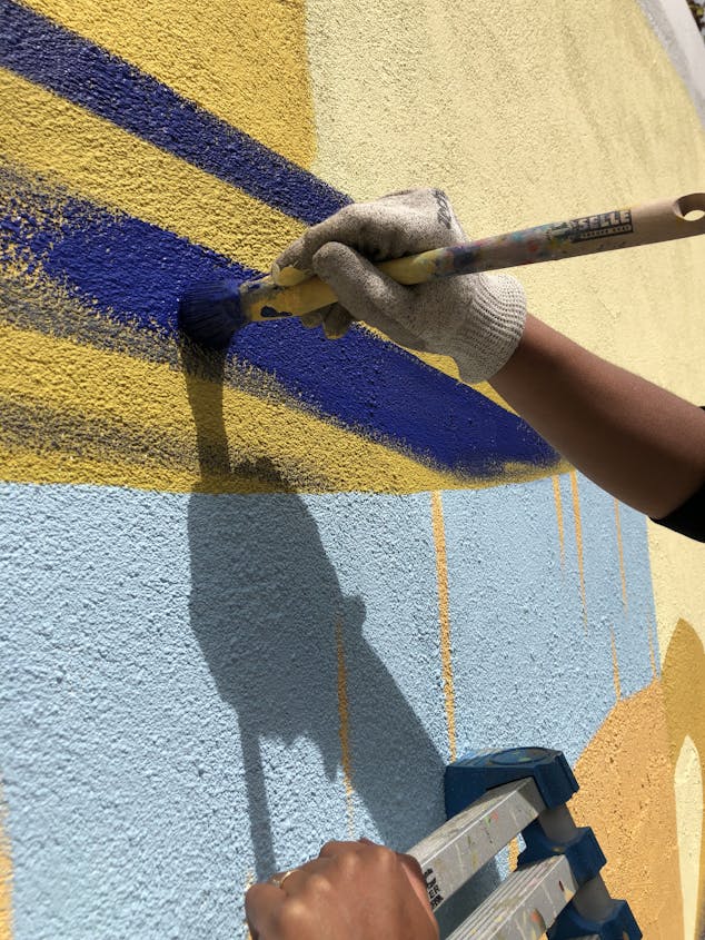 Chantier fresque de la résidence "Les 5 continents". Une main d'artiste effectue un tracé de peinture bleu sur un mur.