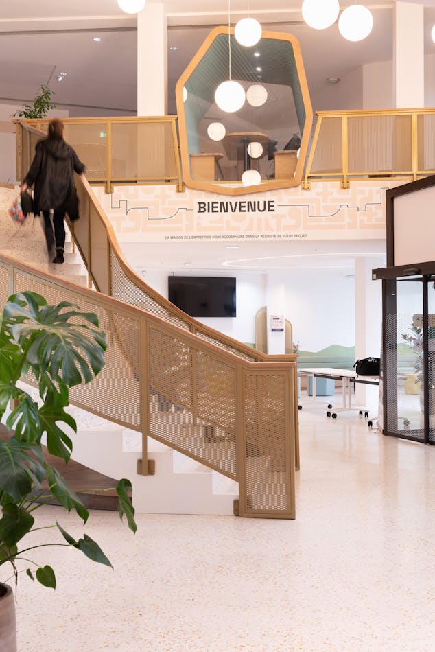 Le hall d'accueil de la nouvelle Maison de l'Entreprise à Saint-Nazaire, dont l'aménagement d'intérieur a été réalisé par Studio Katra. On y voit une personne montant au premier étage grâce aux escaliers.