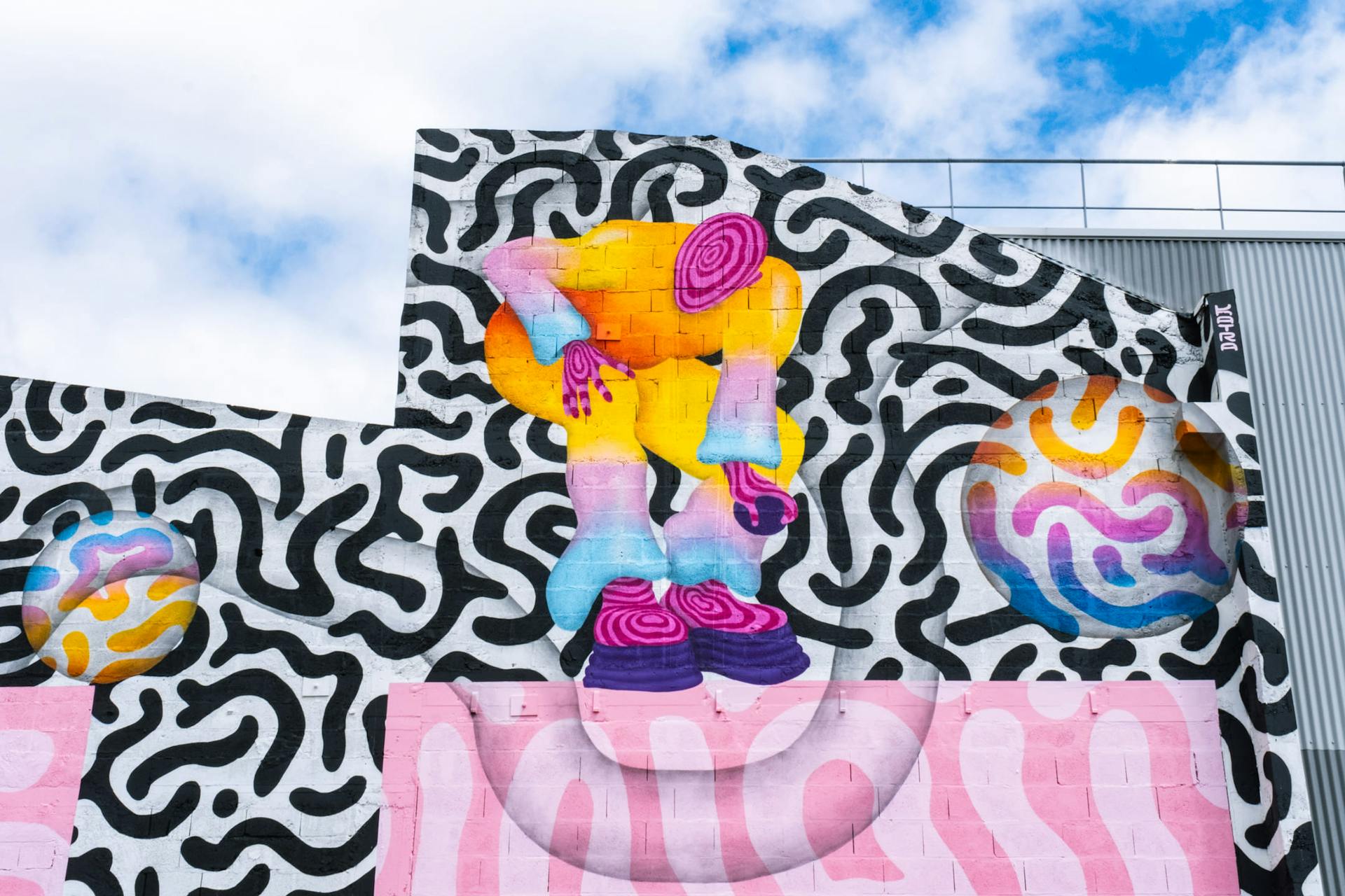 Un espace industriel transformé par une fresque street art de l'artiste Ocü et de l'agence de design Studio Katra, formant un terrain de design actif destiné à la pratique du handball gaélique. On y voit de nombreux motifs psychédéliques aux couleurs vives et un personnage ramassant une balle avec sa main.
