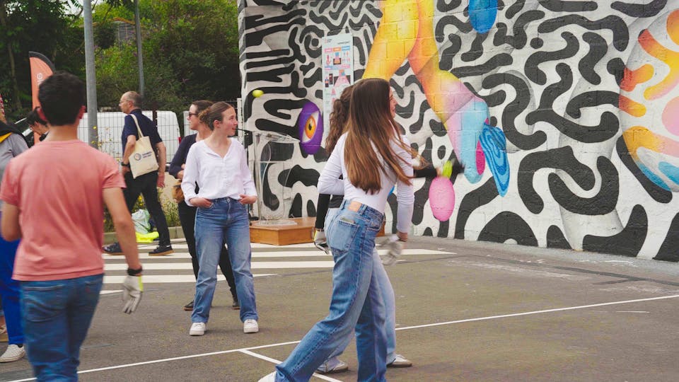 Des joueurs et des joueuses novices s'essayent à la pratique du handball gaélique sur un mur portant une fresque murale street art de l'artiste roumaine Ocü.