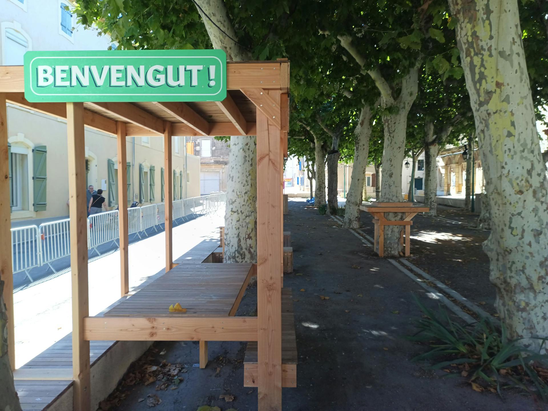 Une terrasse en bois réalisée sur la promenade Marcelin Albert du village d'Argeliers, surmontée d'une enseigne où y est écrit "Bienvenue" en occitan, par l'agence de design Studio Katra