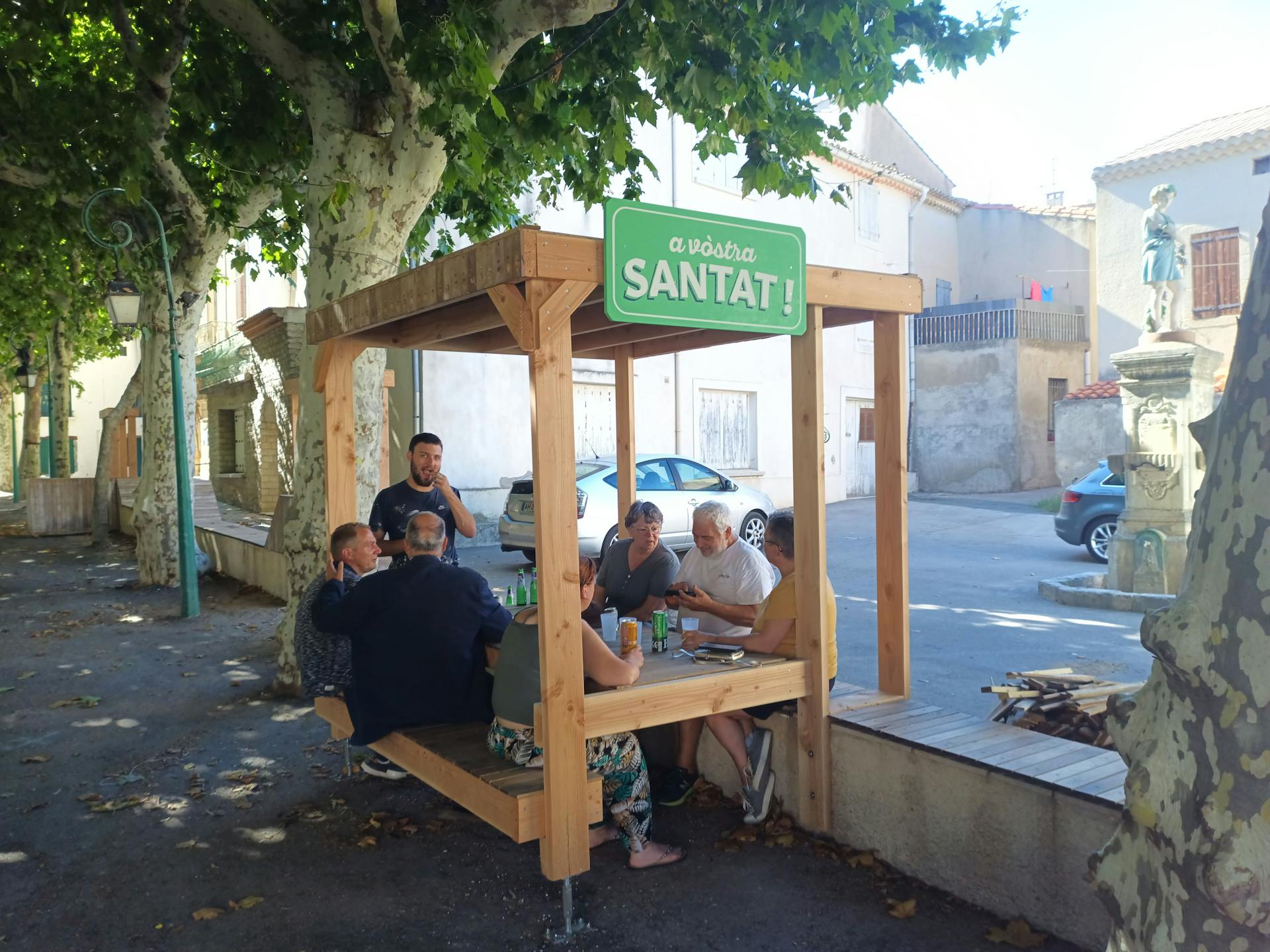 Les habitants d'Argeliers profitent de leur nouvelle terrasse en bois réalisée sur la promenade Marcelin Albert, surmontée d'une enseigne où y est écrit "À votre santé !" en occitan, par l'agence de design Studio Katra