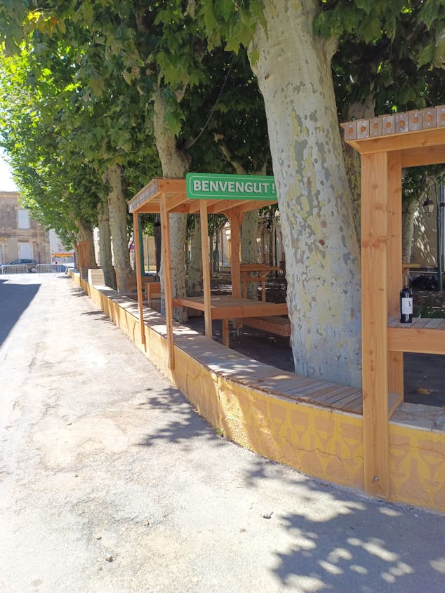 Une terrasse en bois réalisée sur la promenade Marcelin Albert du village d'Argeliers, surmontée d'une enseigne où y est écrit "Bienvenue" en occitan, par l'agence de design Studio Katra