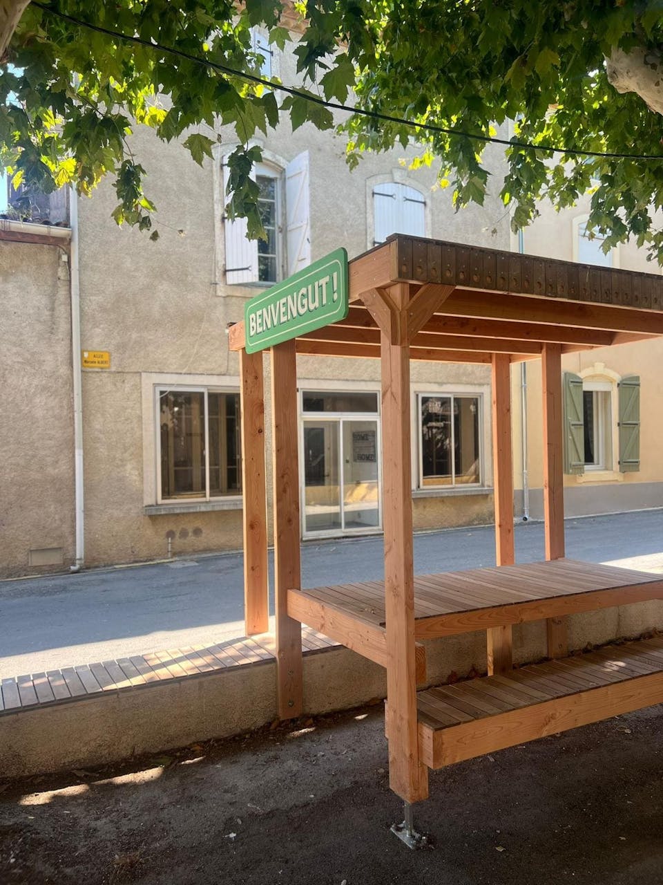 Une terrasse en bois réalisée sur la promenade Marcelin Albert du village d'Argeliers, surmontée d'une enseigne où y est écrit "Bienvenue" en occitan, par l'agence de design Studio Katra