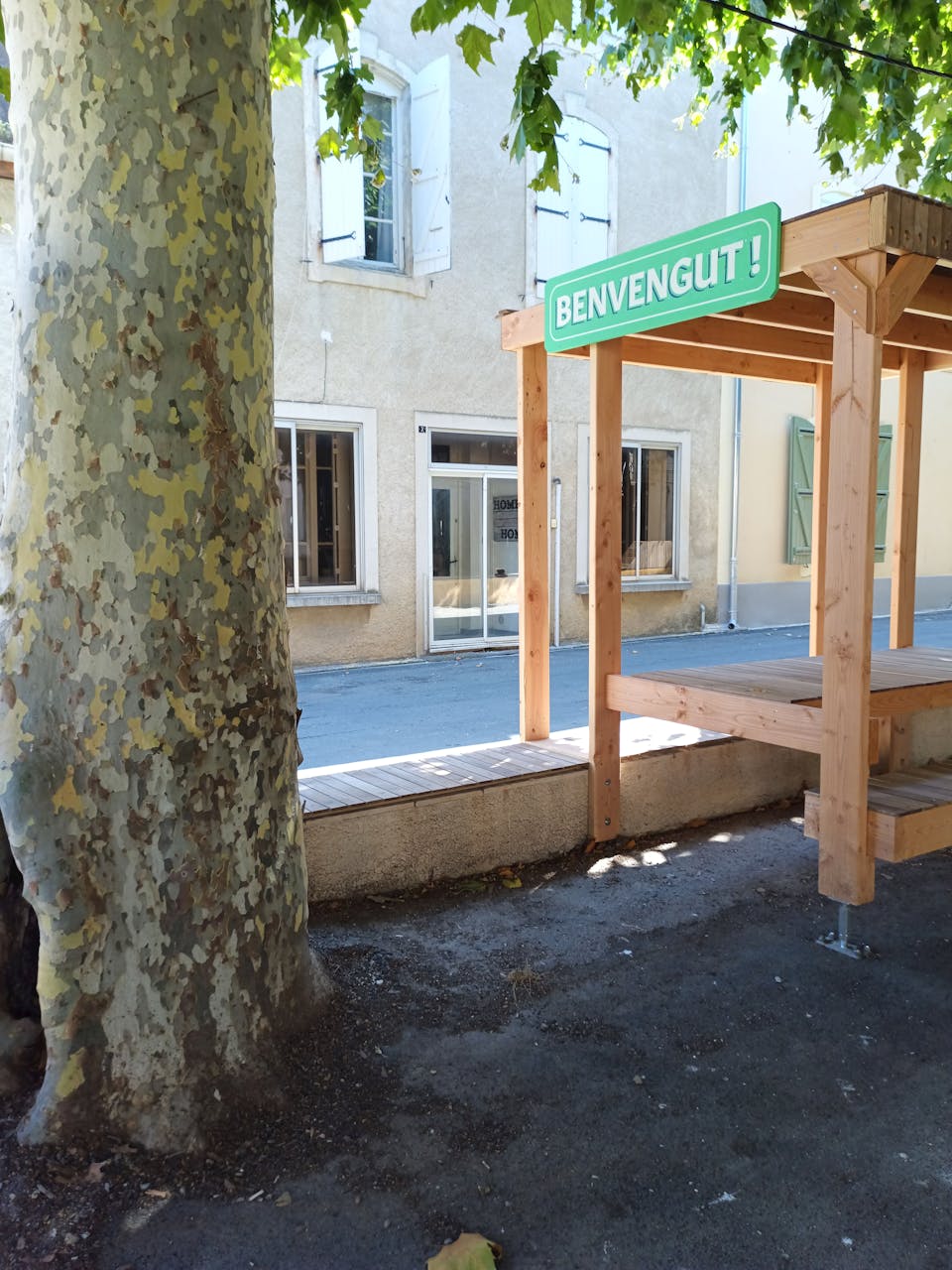 Une terrasse extérieure en bois, habillée d'une enseigne sur laquelle est écrite "Bienvenue !" en occitant, réalisée par l'agence de design Studio Katra.