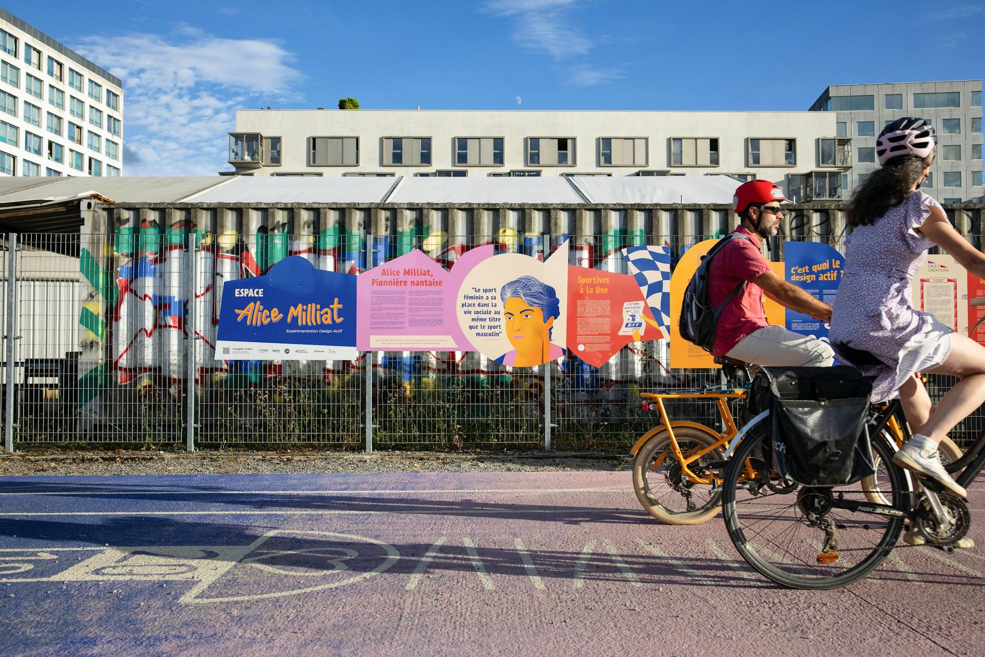 Des panneaux décrivant le nouvel espace Alice Milliat de l'île de Nantes, réalisé par agence de Design Graphique Studio Katra. Des cyclistes passent devant ces panneaux.