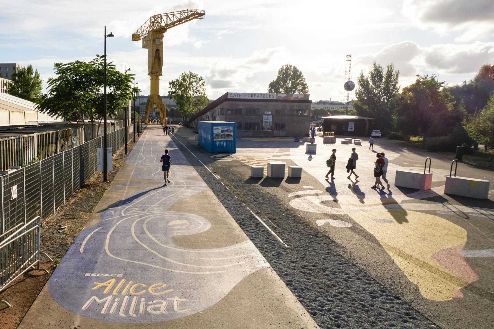 Une fresque peinte sur le sol du nouvel espace de design actif Alice Milliat à Nantes, représentant un personnage féminin en mouvement. Une piste d'athlétisme à l'identité de la fresque est également peinte au sol et liée au projet. Des passants traversent cet espace à pied ou en courant.