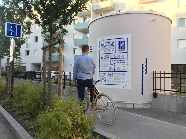 Personne à vélo arrêtée devant une affiche explicative de la nouvelle identité visuelle rue Biesse Nantes.