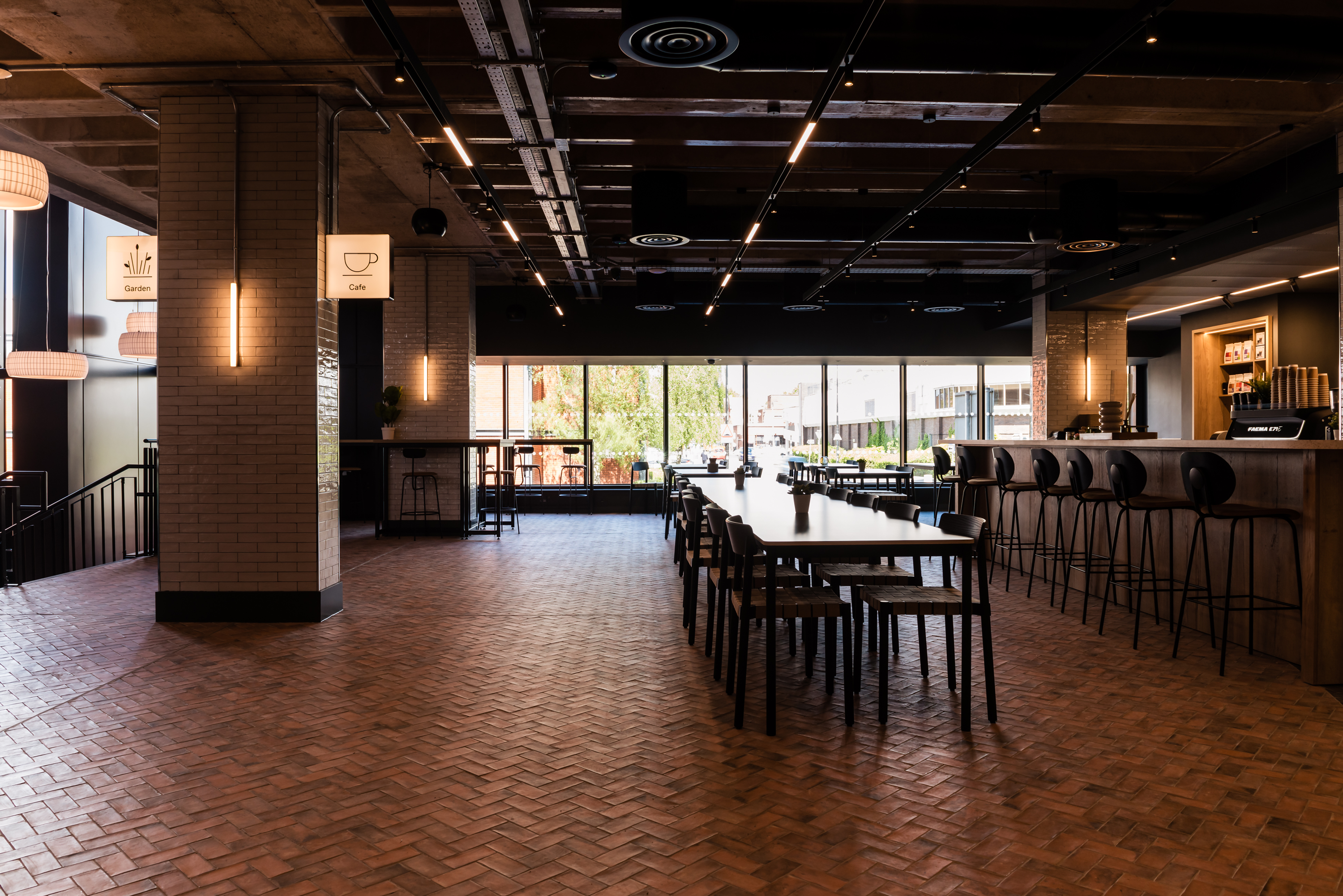 Wide shot of the cafe, showing a long table with chairs, bar seating, parquet floor and tiled pillars.