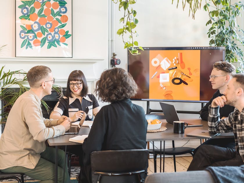 The team in discussion around a table, in a room with hanging plants, a bold print and TV showing a colourful illustration.