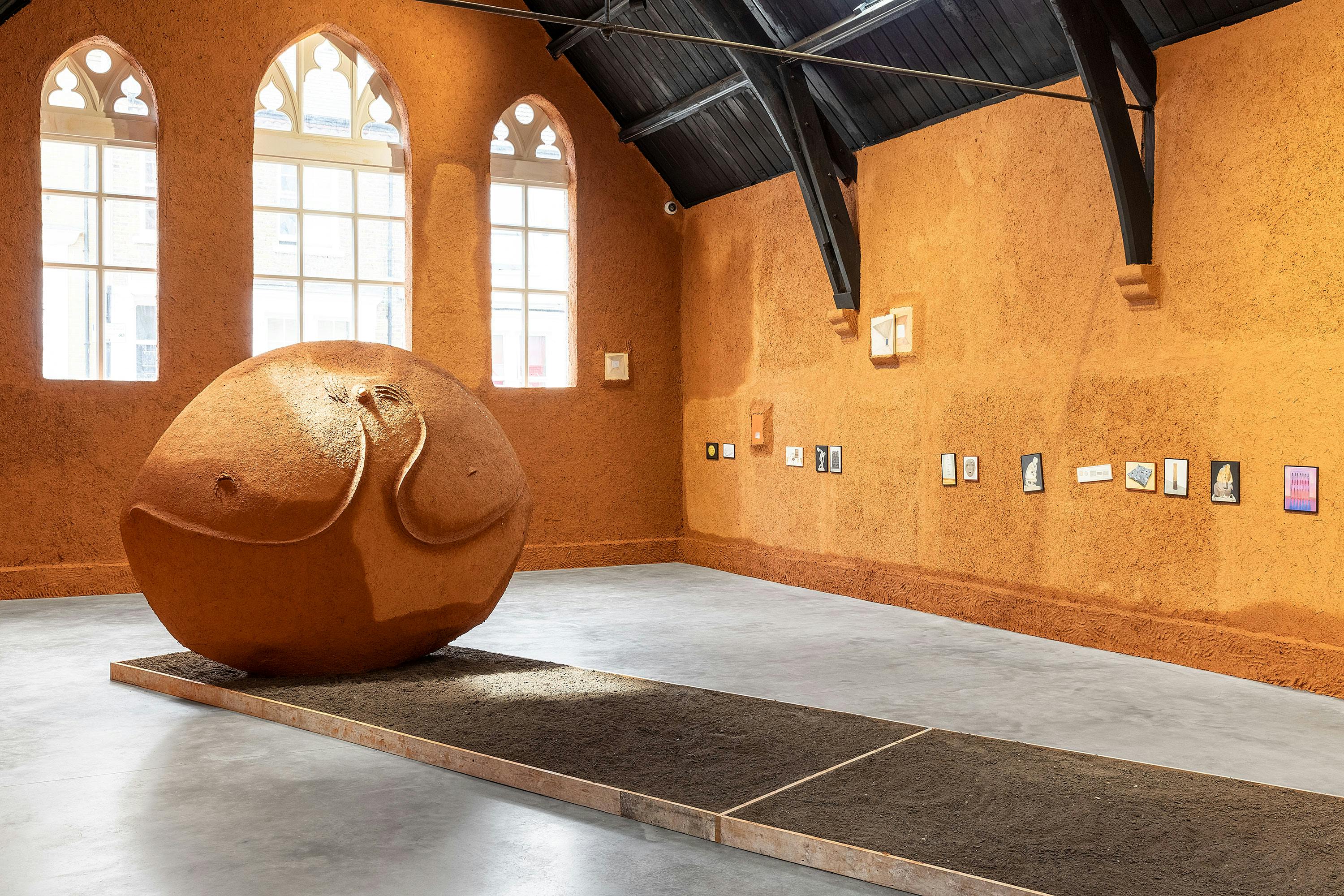 A gallery with walls entirely covered in terracotta coloured mud clay, with a vaulted black ceiling. In the centre of a room stands a large round sculpture made of mud clay, sitting in a raised bed of brown earth. Small paintings of publicly held artworks are arranged on the walls