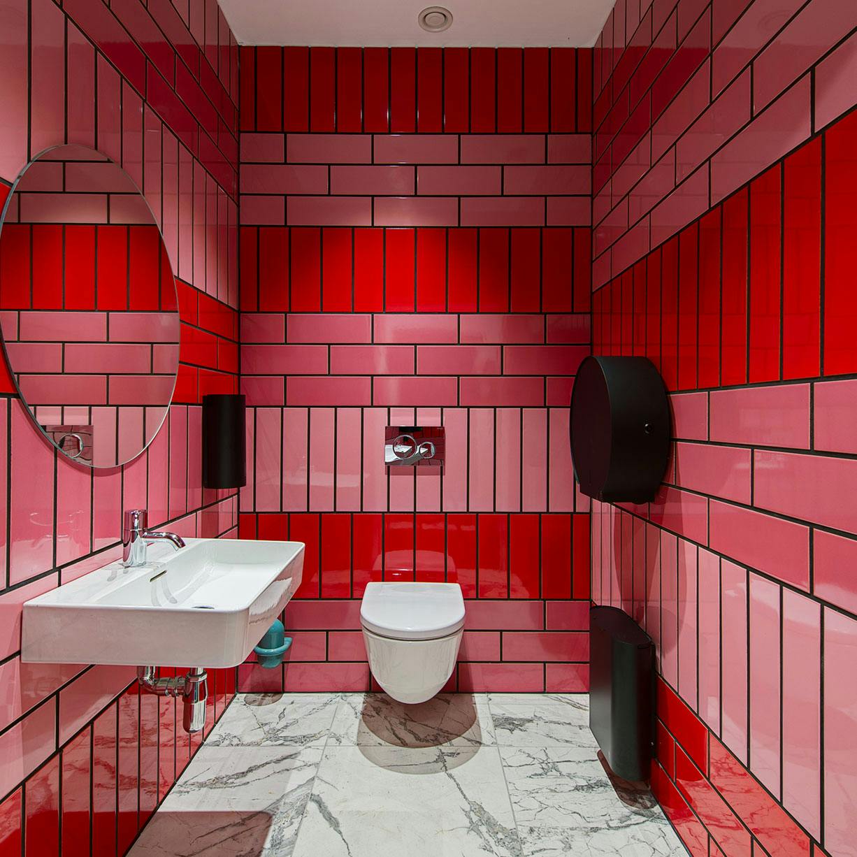 A modern bathroom with red and pink tiled walls, a white floating sink with a round mirror above, and a wall-mounted toilet. The floor has white and grey marble tiles. Black fixtures and accessories contrast against the vibrant tiles.