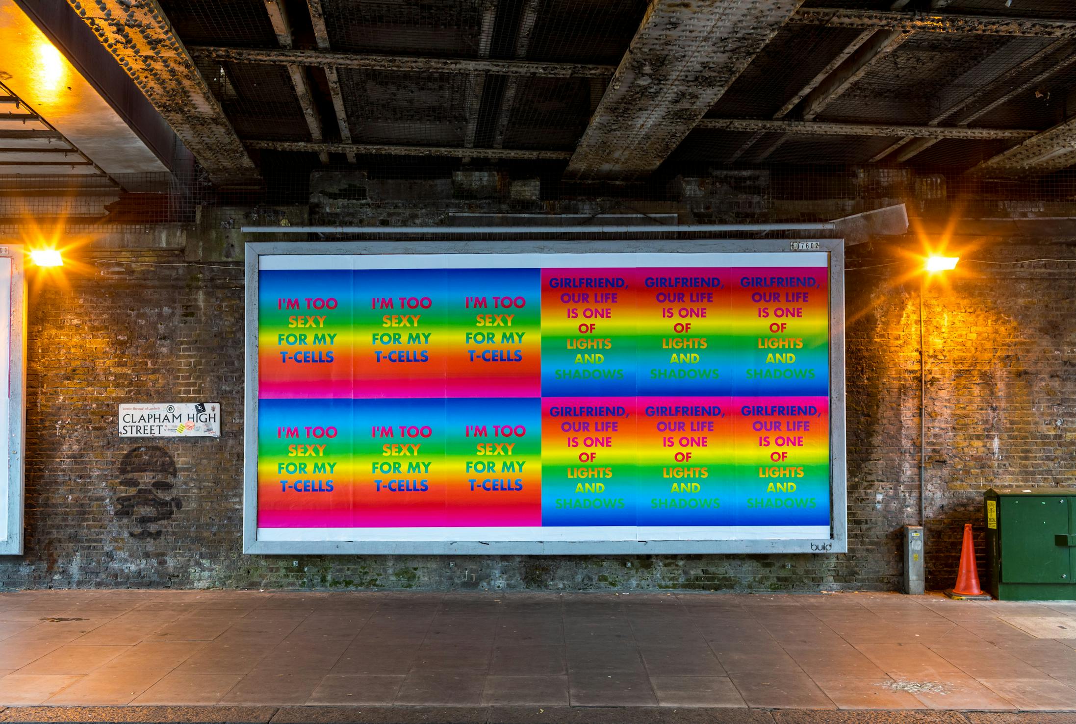 A colourful billboard under a bridge displays repeating posters by David McDiarmid with rainbow text blocks featuring phrases like "I'M TOO SEXY FOR MY T-CELLS" and "OUR LIFE IS ONE OF LIGHTS AND SHADOWS" on a brick wall at Clapham High Street.
