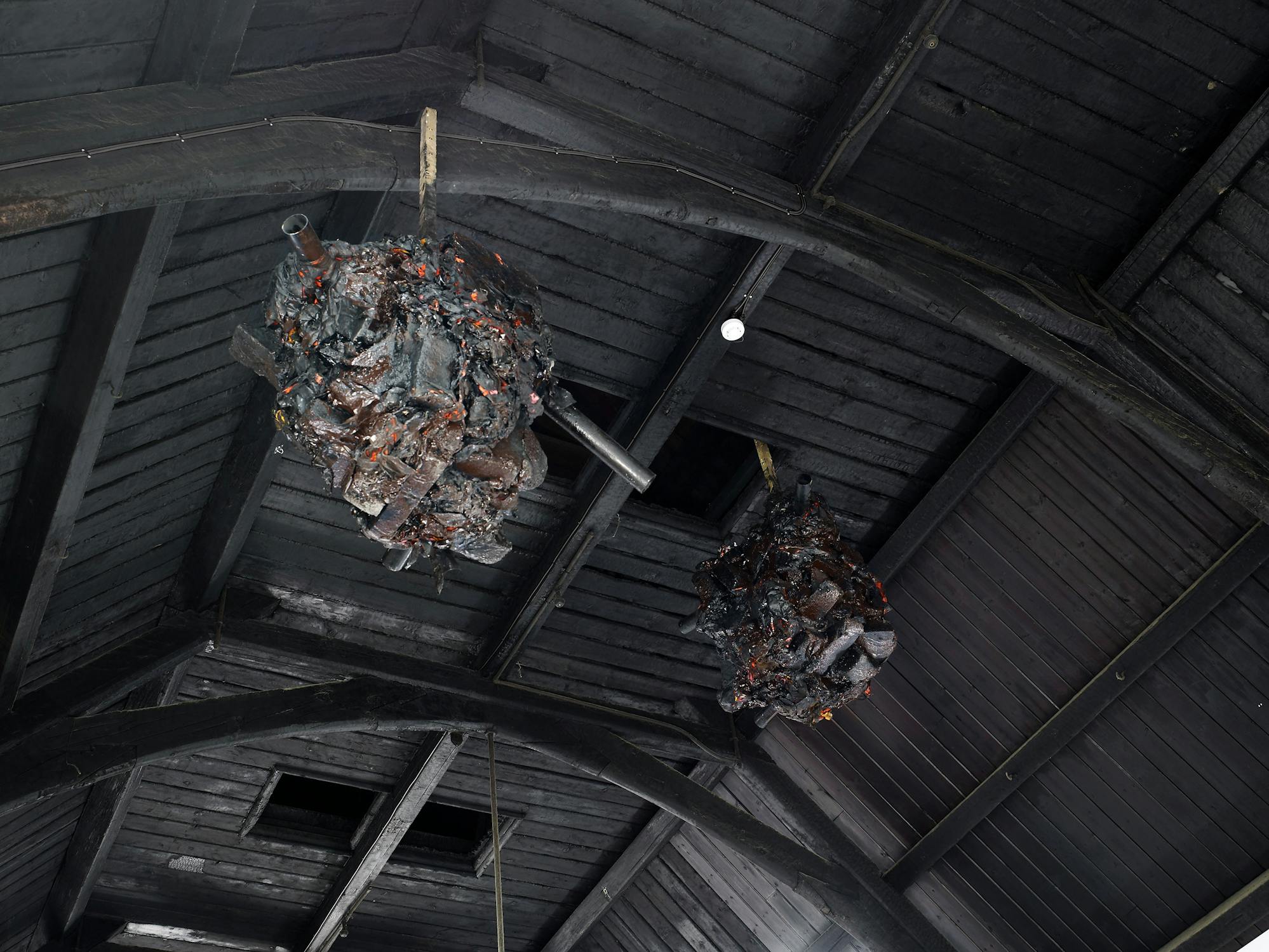 Two large, dark, spherical sculptures hang from the wooden rafters of a high ceiling. The sculptures appear burnt, with glowing red embers scattered throughout, resembling smouldering debris or nests within a dimly lit, rustic interior.