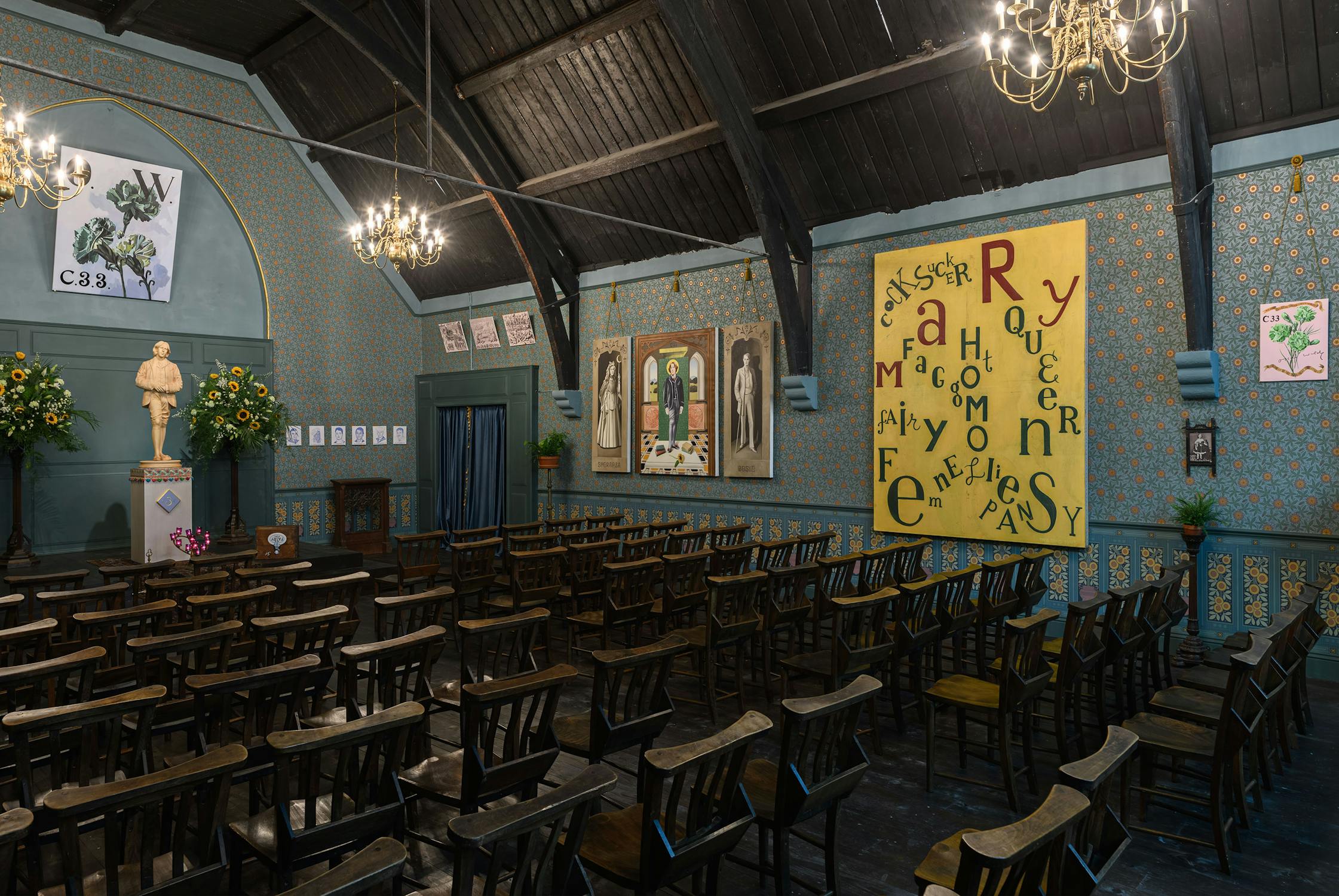 An historic room with wooden chairs facing a small stage adorned with a statue and floral arrangements. The walls are decorated with blue patterned wallpaper, chandeliers, framed art, and a large yellow board with jumbled letters in black and green.