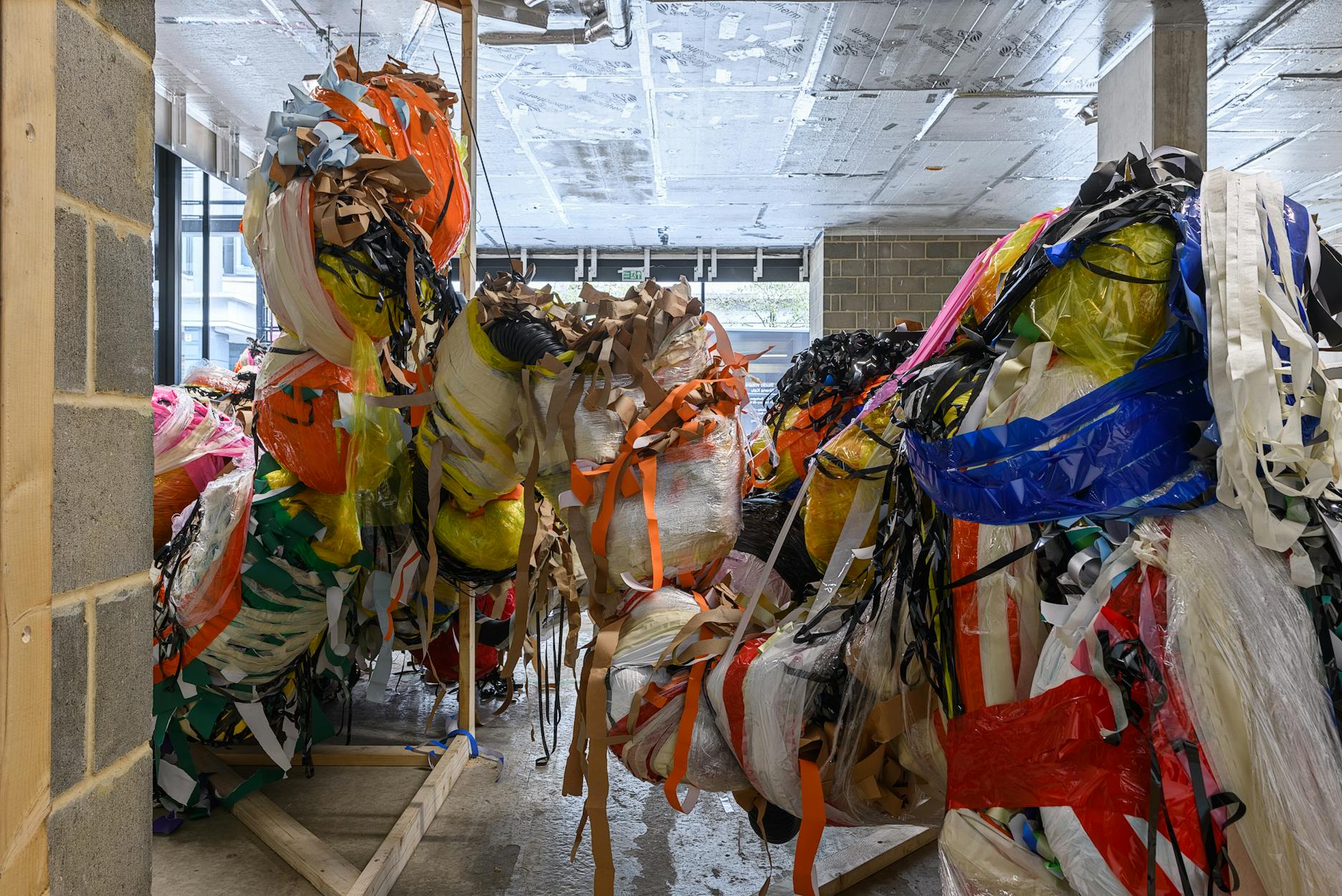 Large, colourful bundles of crumpled plastic sheets, fabric strips, and ribbons are tied together and suspended in an unfinished room with cement floors, exposed pipes, and brick walls. Natural light comes through the window in the background.