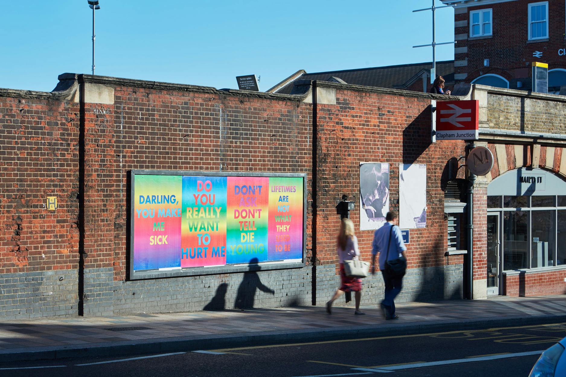 Two people walk past a brick wall with colourful text art reading, "DARLING, YOU MAKE ME SICK. DO YOU REALLY WANT TO HURT ME? DON'T DIE YET. LIFETIMES ARE WHAT THEY DUST. DON'T WAIT TILL DIE TO LOVE.