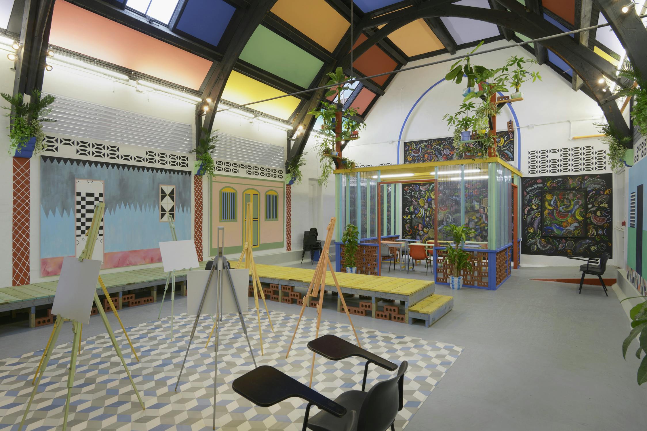 Sol Calero’s installation at Studio Voltaire, depicting a large, vividly coloured studio space with high ceilings, tiled flooring, easels, contemporary furniture, and lush potted plants. Sunlight filters through from above, illuminating the mural-covered walls.