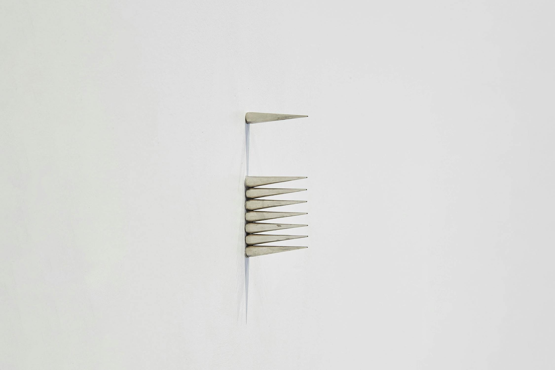 Minimalist artwork by Solomon Garçon featuring eight metallic, cone-shaped spikes protruding horizontally from a white wall, arranged in a vertical line with the points facing right. The overall composition is simple and clean.