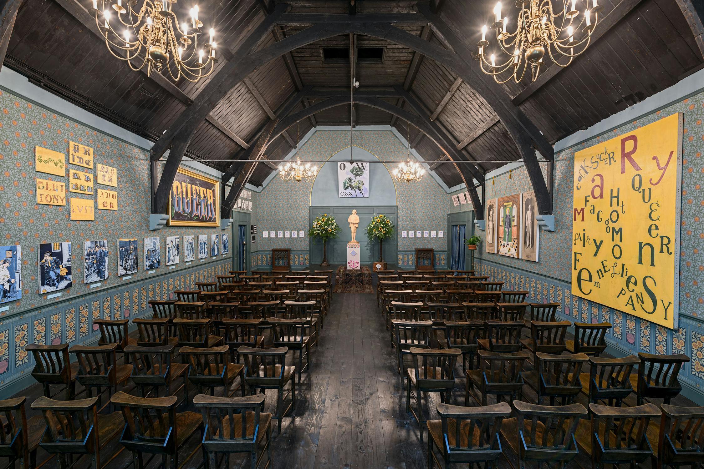 A vintage hall with wooden chairs arranged in rows, ornate chandeliers, dark wooden ceiling, blue and gold patterned walls, large colourful posters, and framed photographs. An altar with flowers and portrait is at the front, evoking an old-fashioned gathering space.
