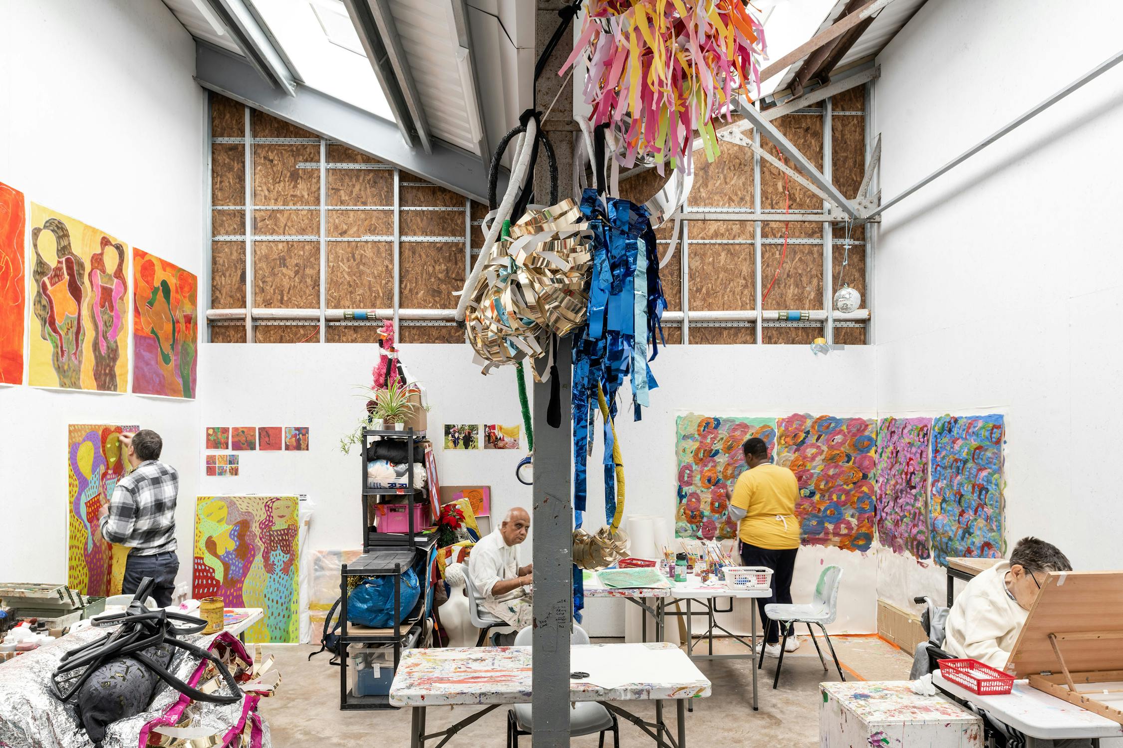 A colorful art studio with vibrant paintings on the walls. Three people are engaged in various creative activities at tables. The room is decorated with hanging ribbons and artwork, under a ceiling with skylights.