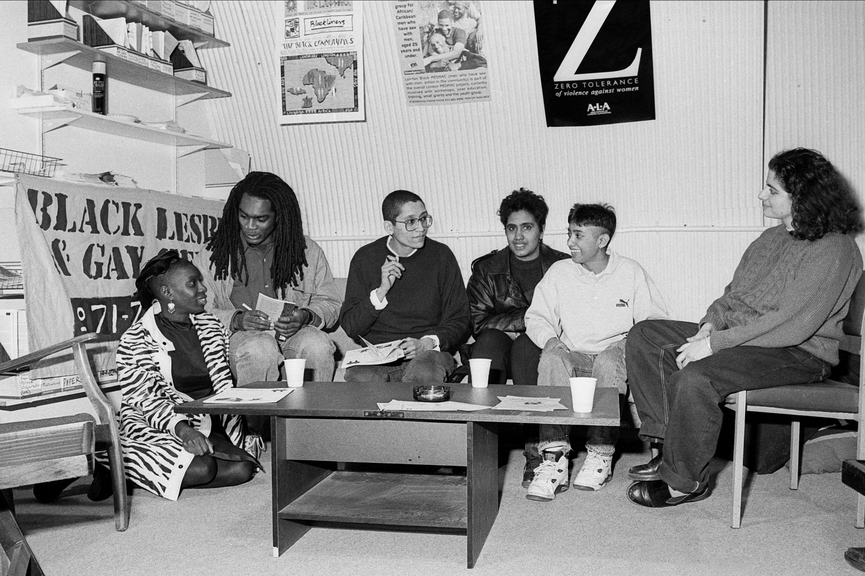 A black and white photograph of group of young Black and Asian people sitting speaking. A banner for the Black Lesbian and Gay Centre is in the background.