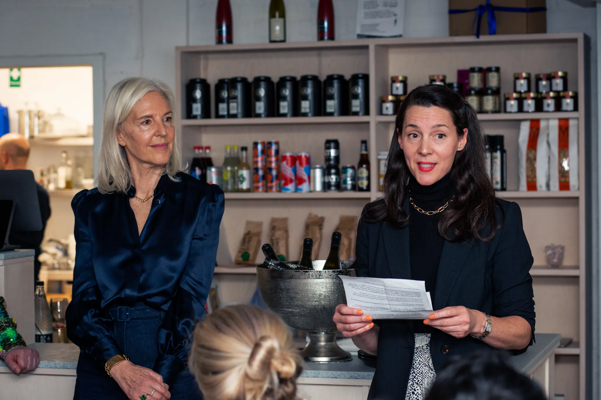 Two women stand in front of shelves with drinks and wine bottles. Victoria Sidall, who has long dark hair and a black blazer speaks while holding papers. The other, with blonde hair and a navy blouse, listens attentively. People sit facing them in the foreground.