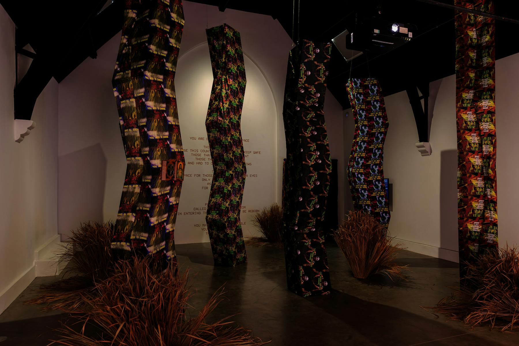 A photograph of a dimly lit room, that is filled with tall angular structures that reach from the floor to the ceiling. The structures are wrapped in vibrantly patterned fabrics. a wall in the background is downlit and features a set of instructions in red letters.