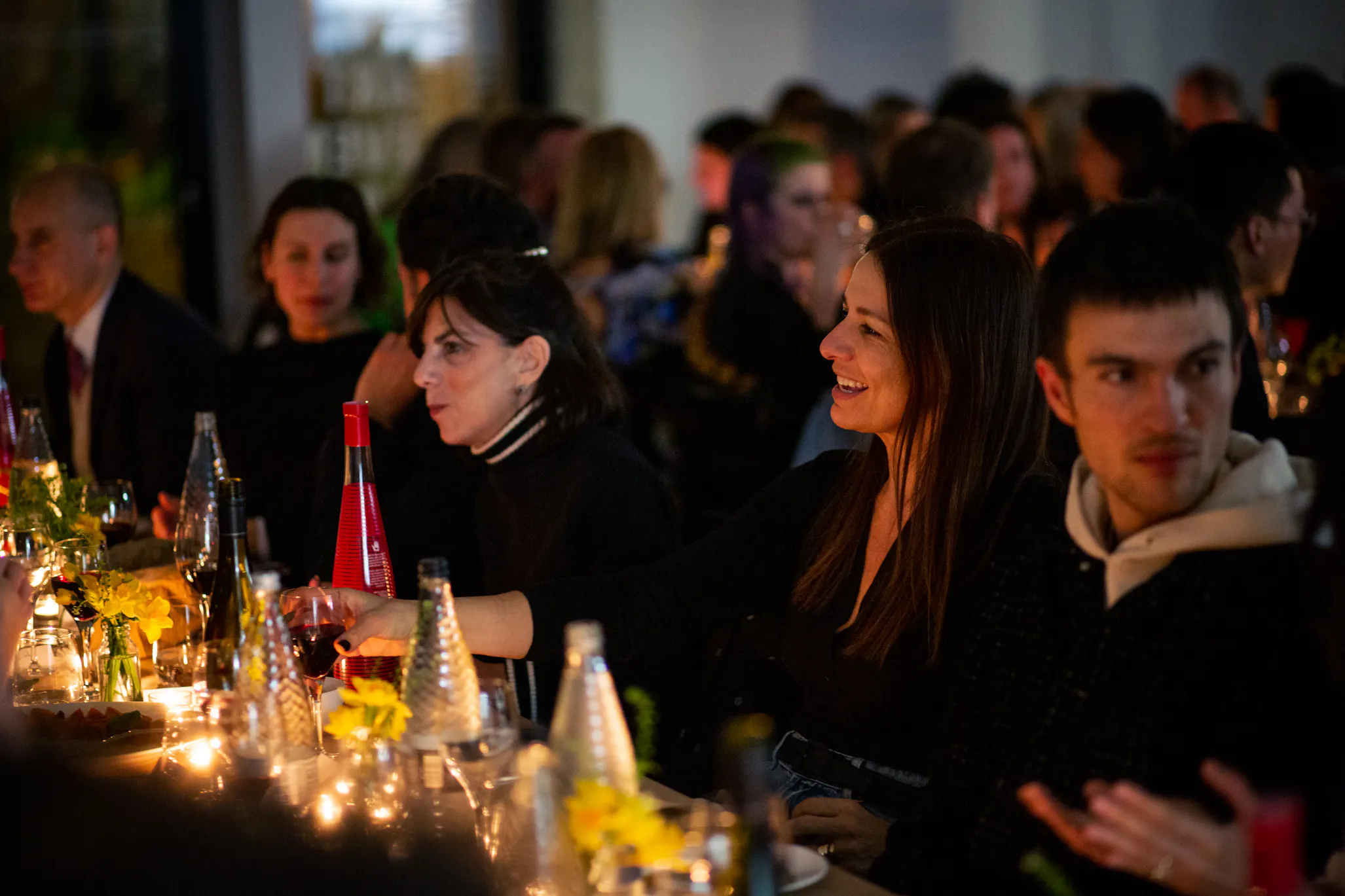A warmly lit dinner scene with people seated at a long table. Glass bottles, yellow flowers, and candles decorate the table. A woman in the centre smiles and reaches across the table, surrounded by others engaged in conversation.